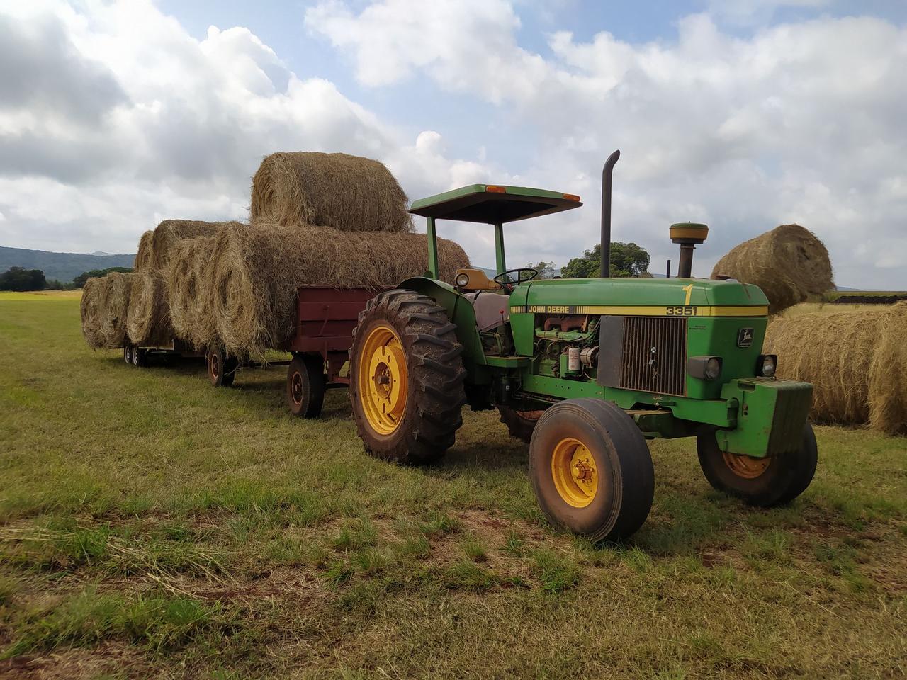 A green tractor pulling a trailer loaded with large hay bales in a rural field.
