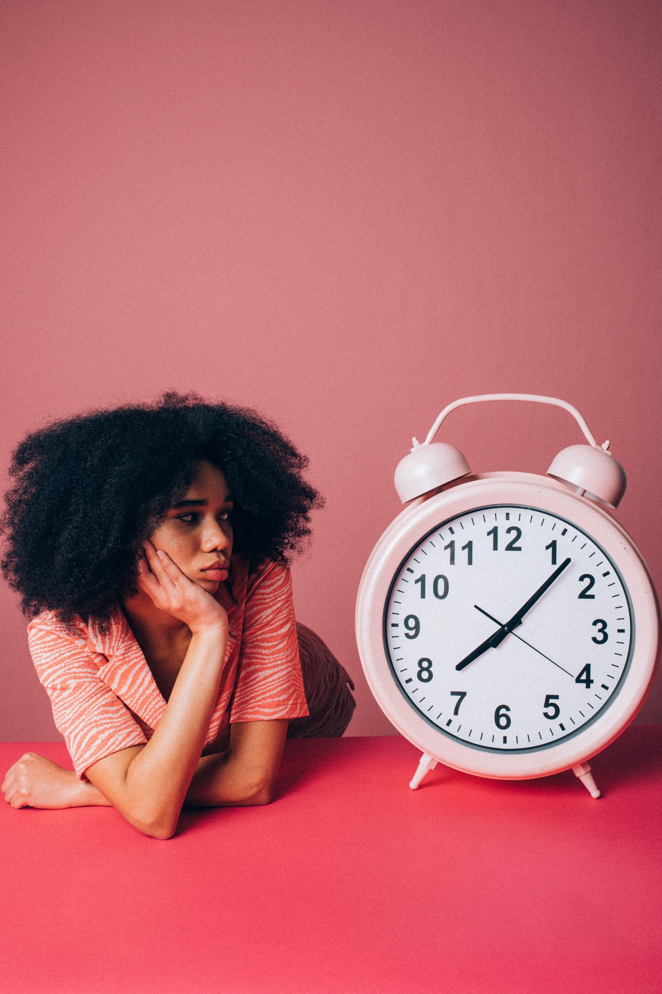 Woman in a pink shirt leans on table with a large alarm clock, conveying impatience.