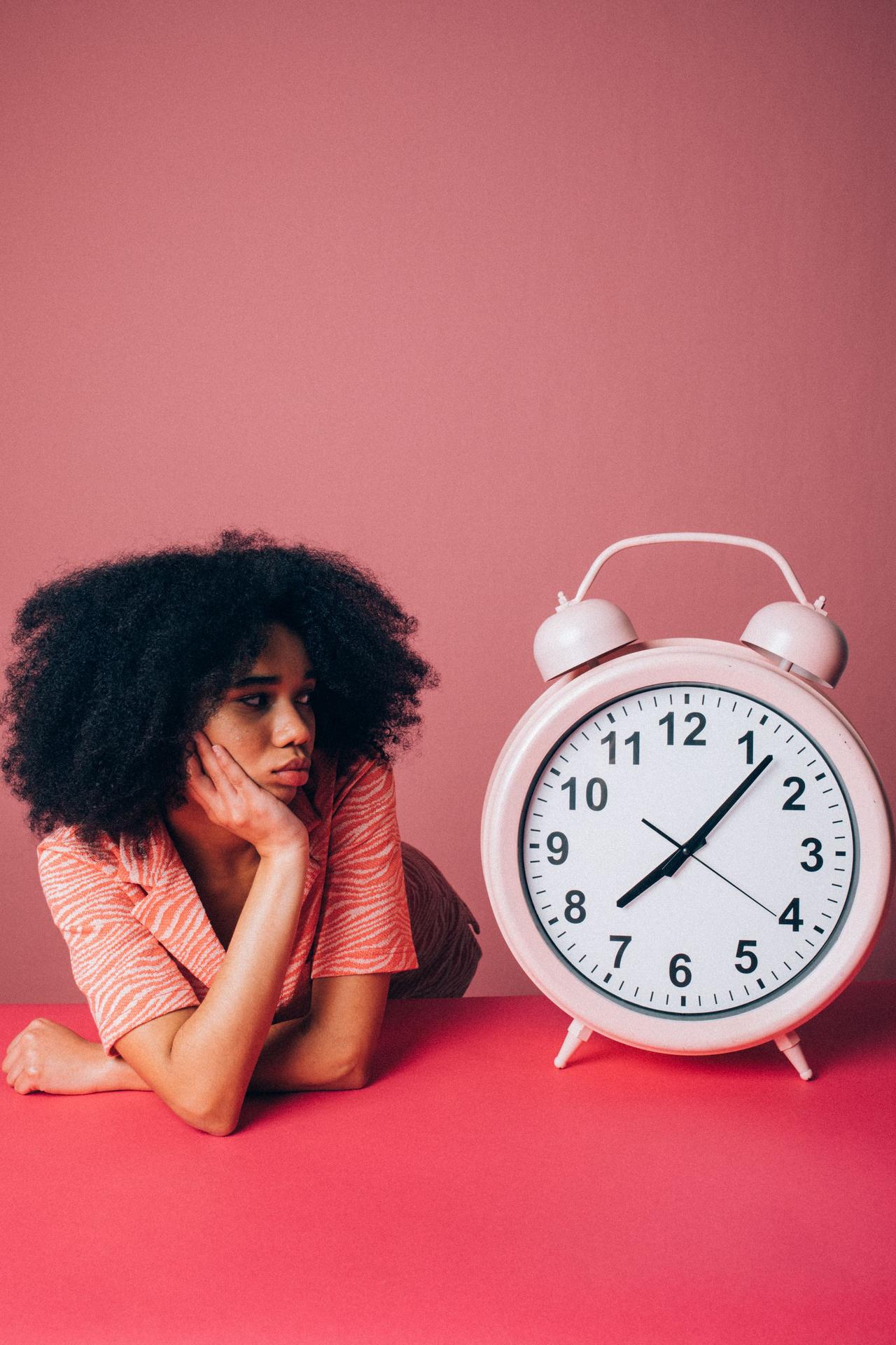 Woman in a pink shirt leans on table with a large alarm clock, conveying impatience.