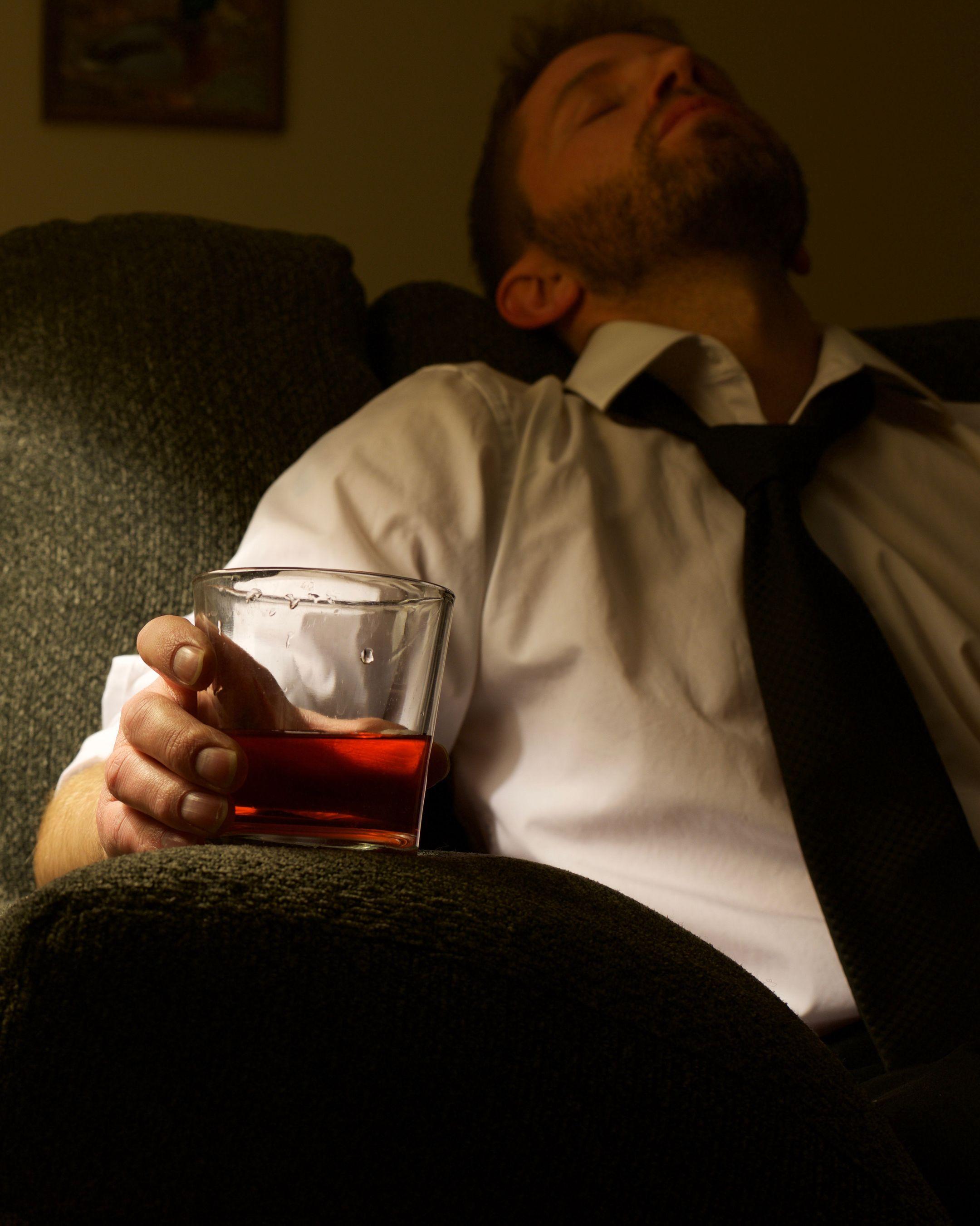 A man in a white shirt and tie relaxes on a couch with a glass of whisky.