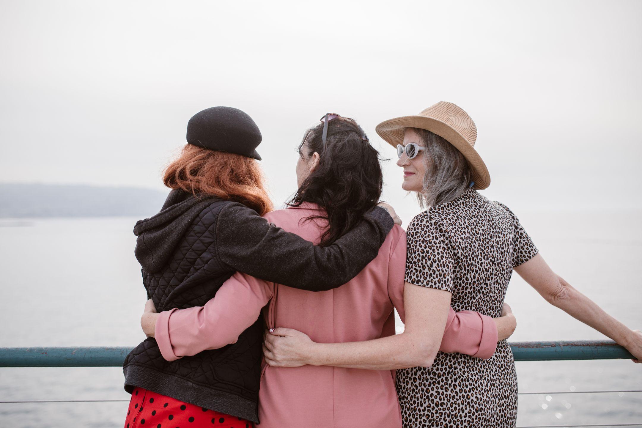 Three women, seen from behind, embracing by a seaside railing, symbolizing friendship.