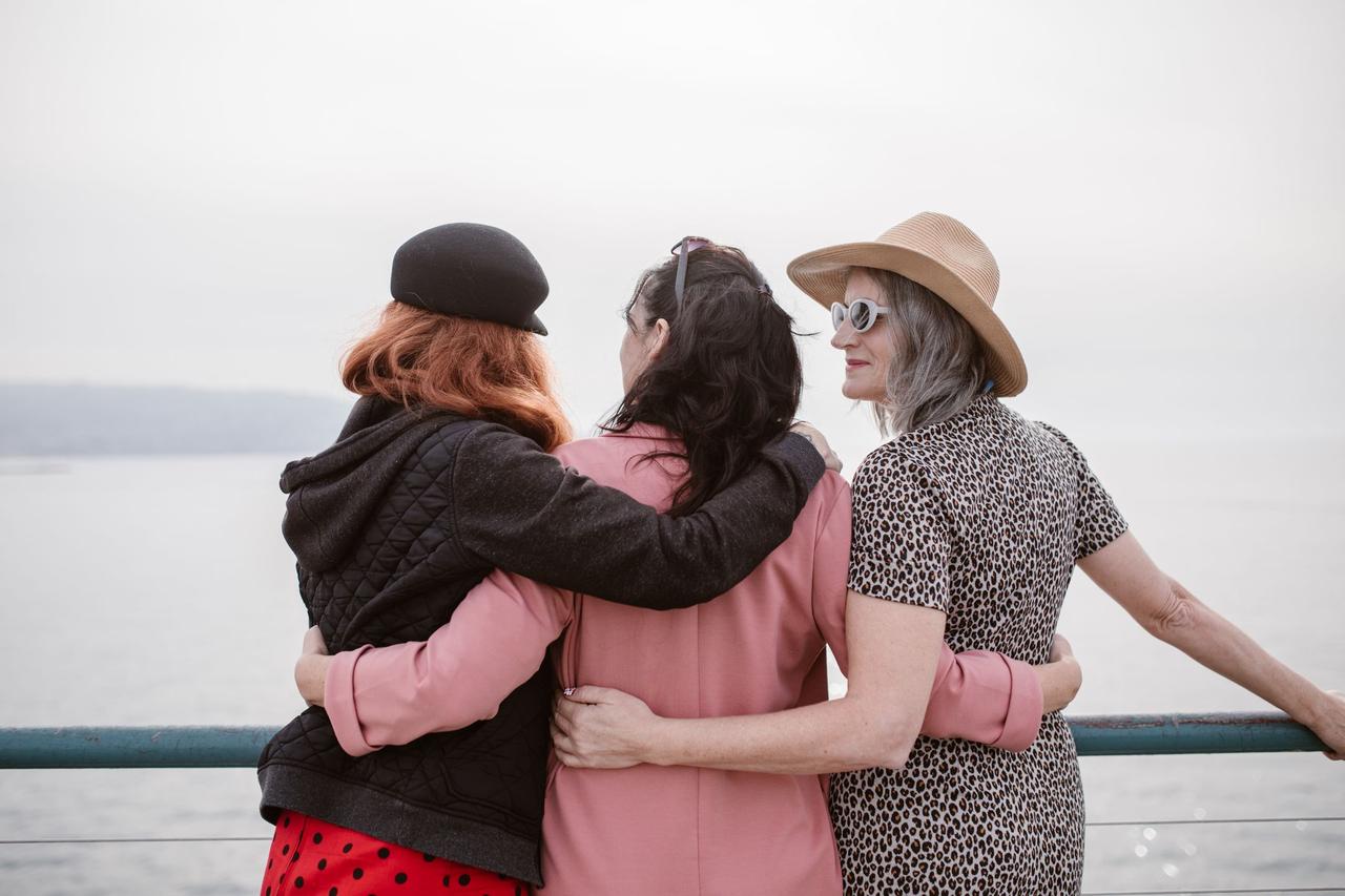 Three women, seen from behind, embracing by a seaside railing, symbolizing friendship.