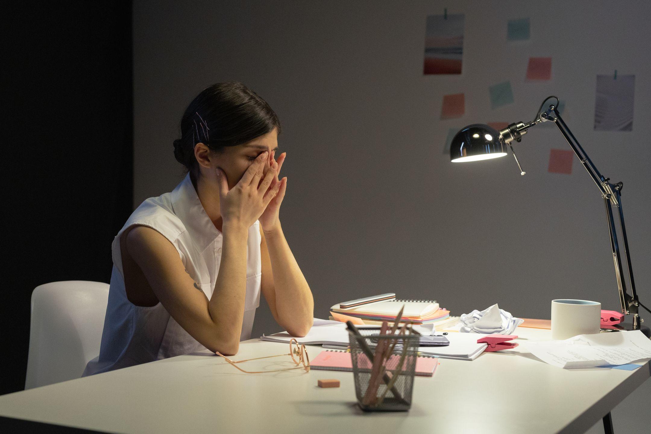 A stressed businesswoman rubs her eyes at a cluttered desk, overwhelmed by work.