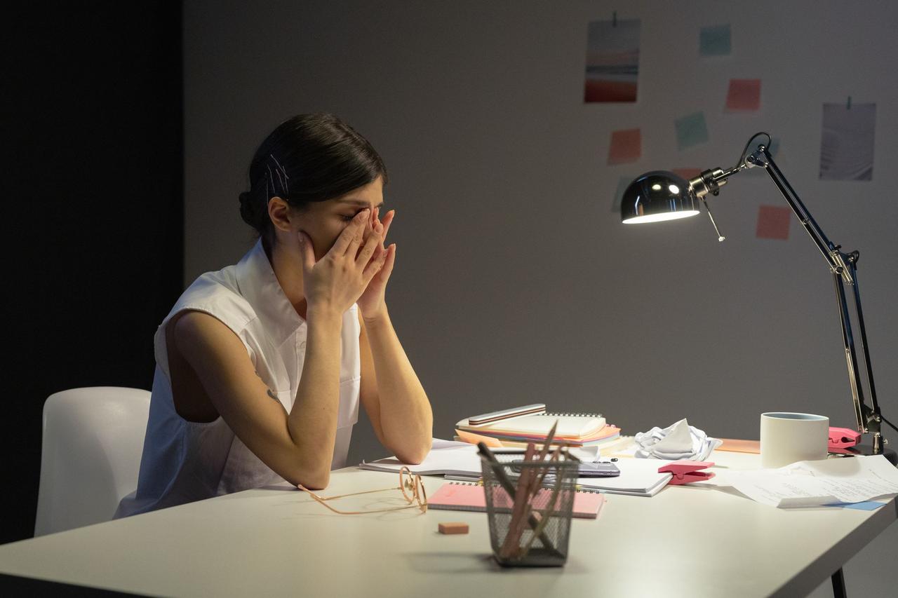 A stressed businesswoman rubs her eyes at a cluttered desk, overwhelmed by work.
