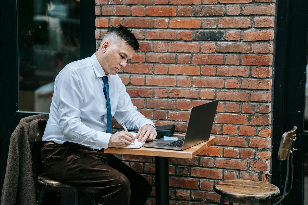 Adult man in corporate attire working on laptop at a cafe table outdoors.