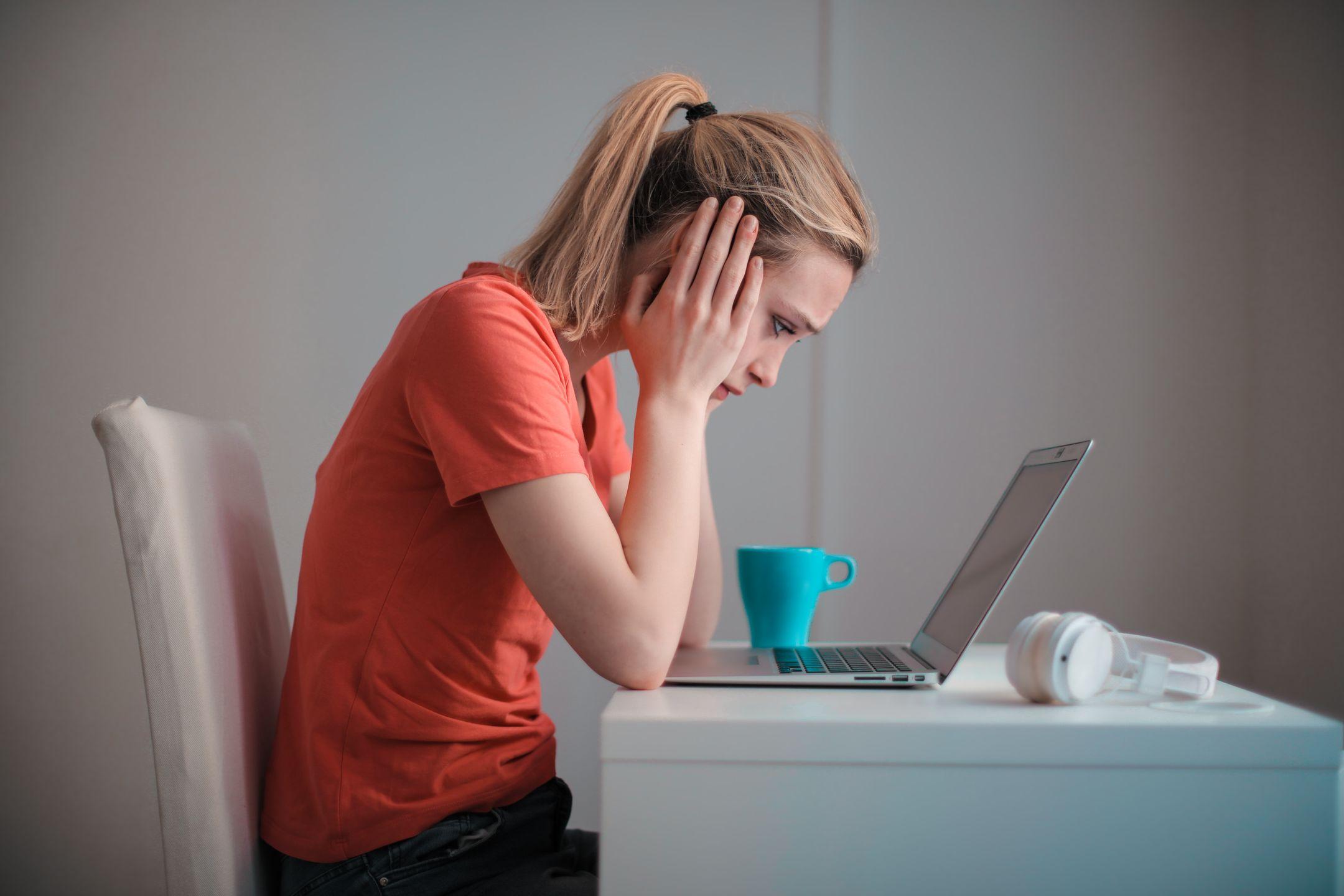 Side view of thoughtful worried female student in casual clothes focusing on screen and trying to solve problem while sitting at table with blue mug of hot drink and headphones and working on laptop at home