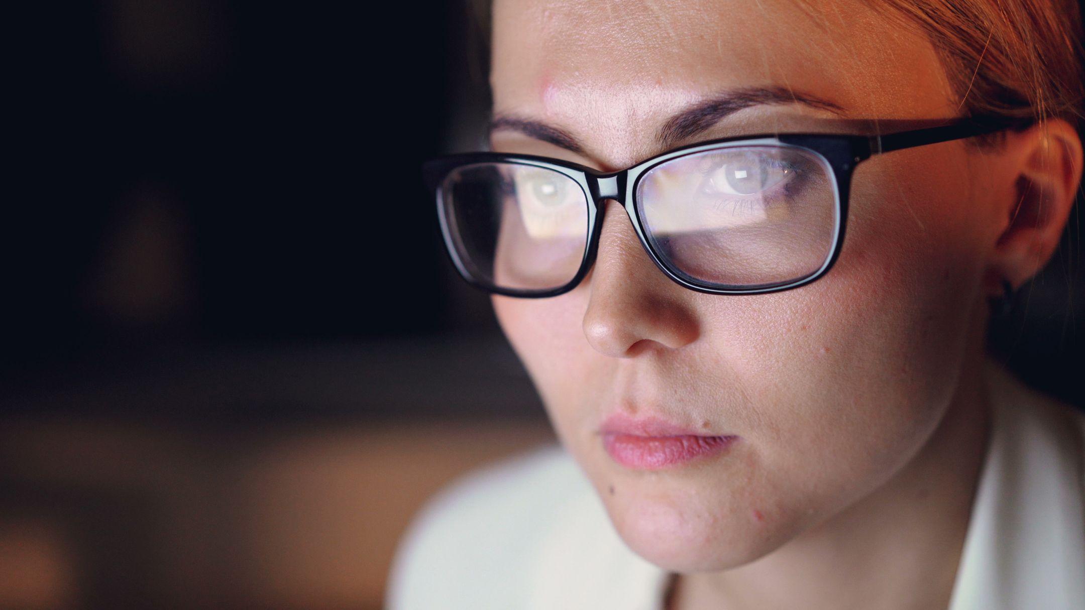 Close-up of a woman concentrating on a screen, wearing glasses and working late at night.