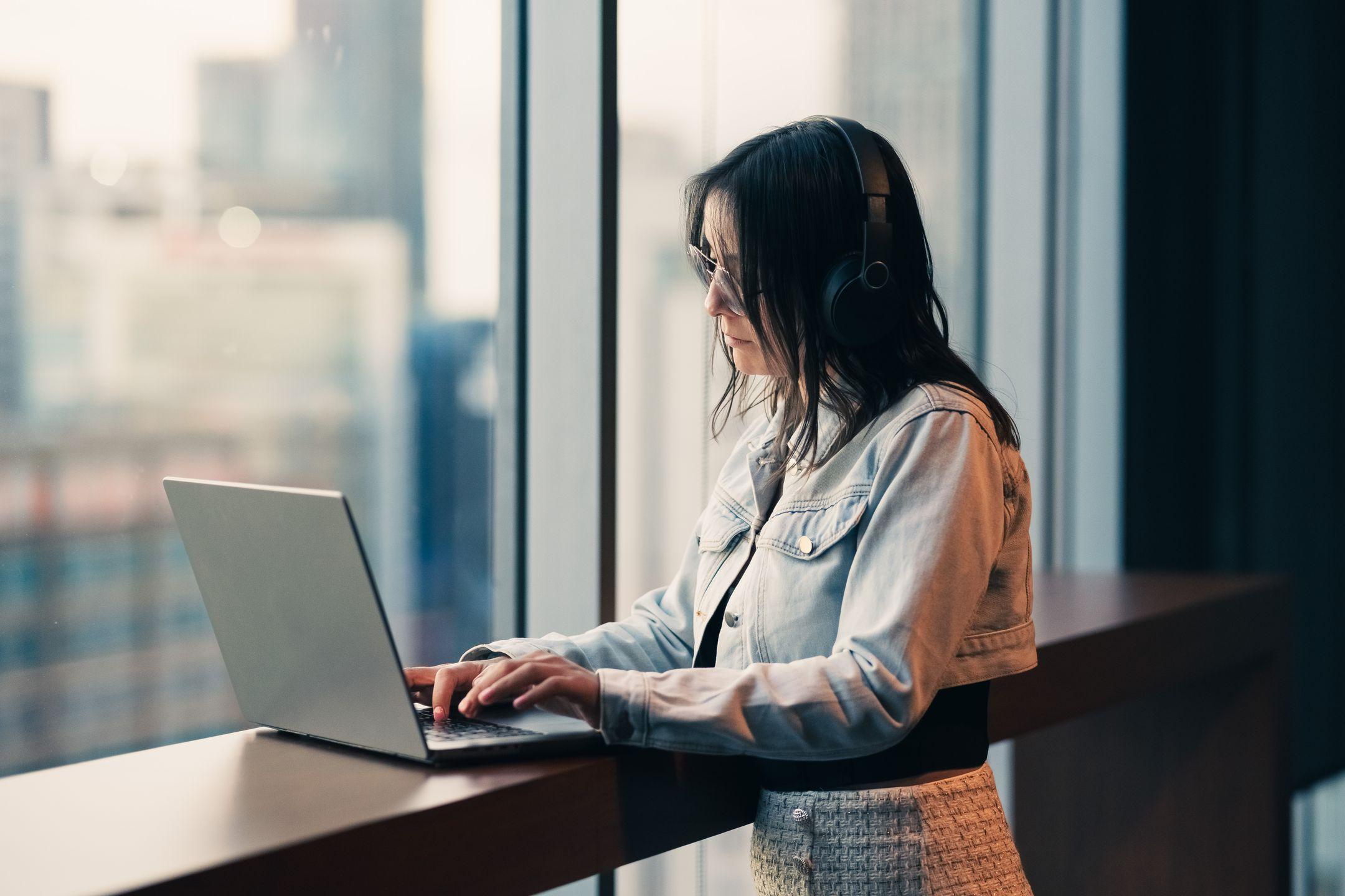 A woman wearing headphones works on her laptop in a high-rise building, looking out a large window with a city view.