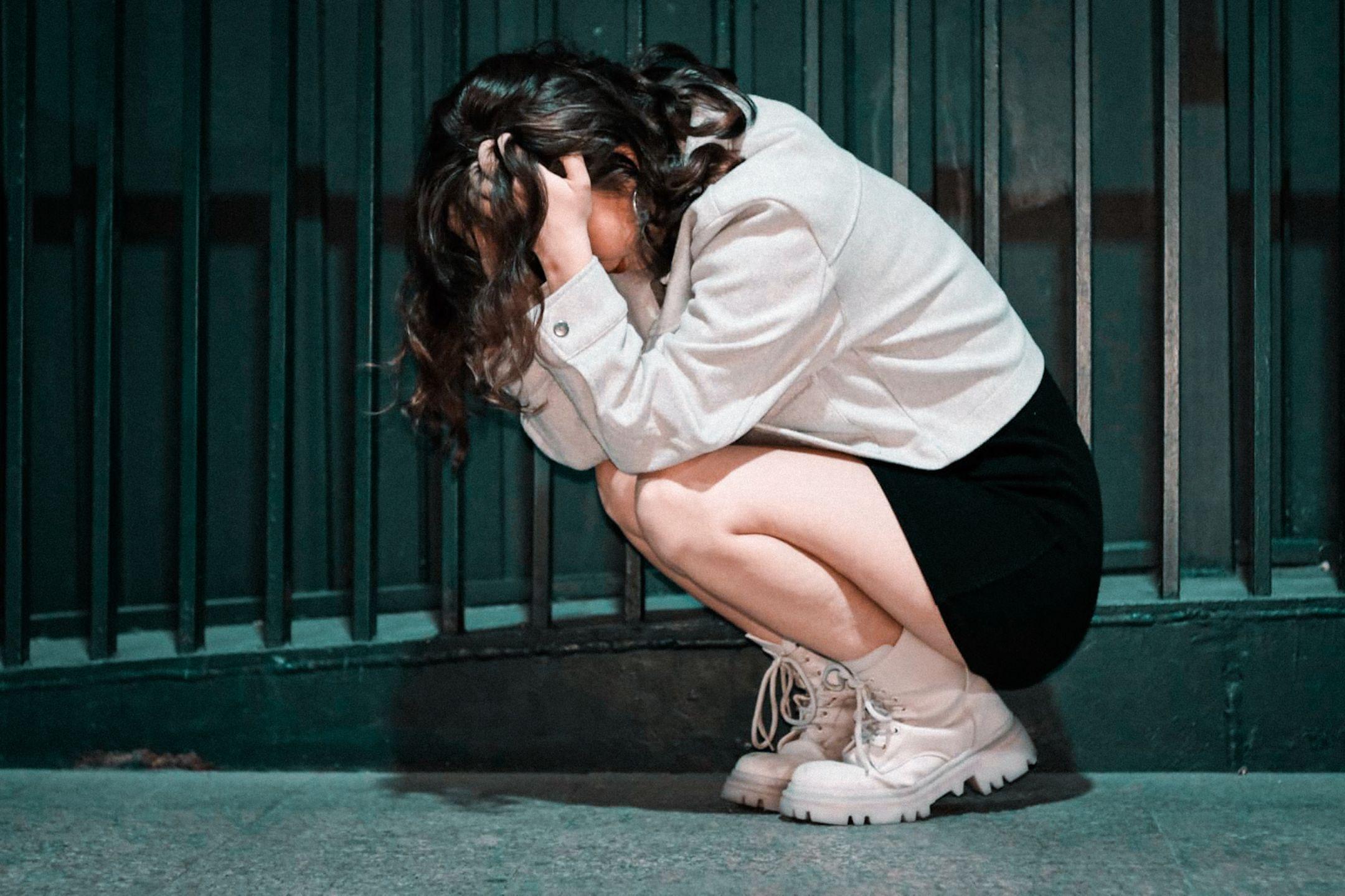 Woman crouching with head in hands against city wall, expressing deep contemplation.