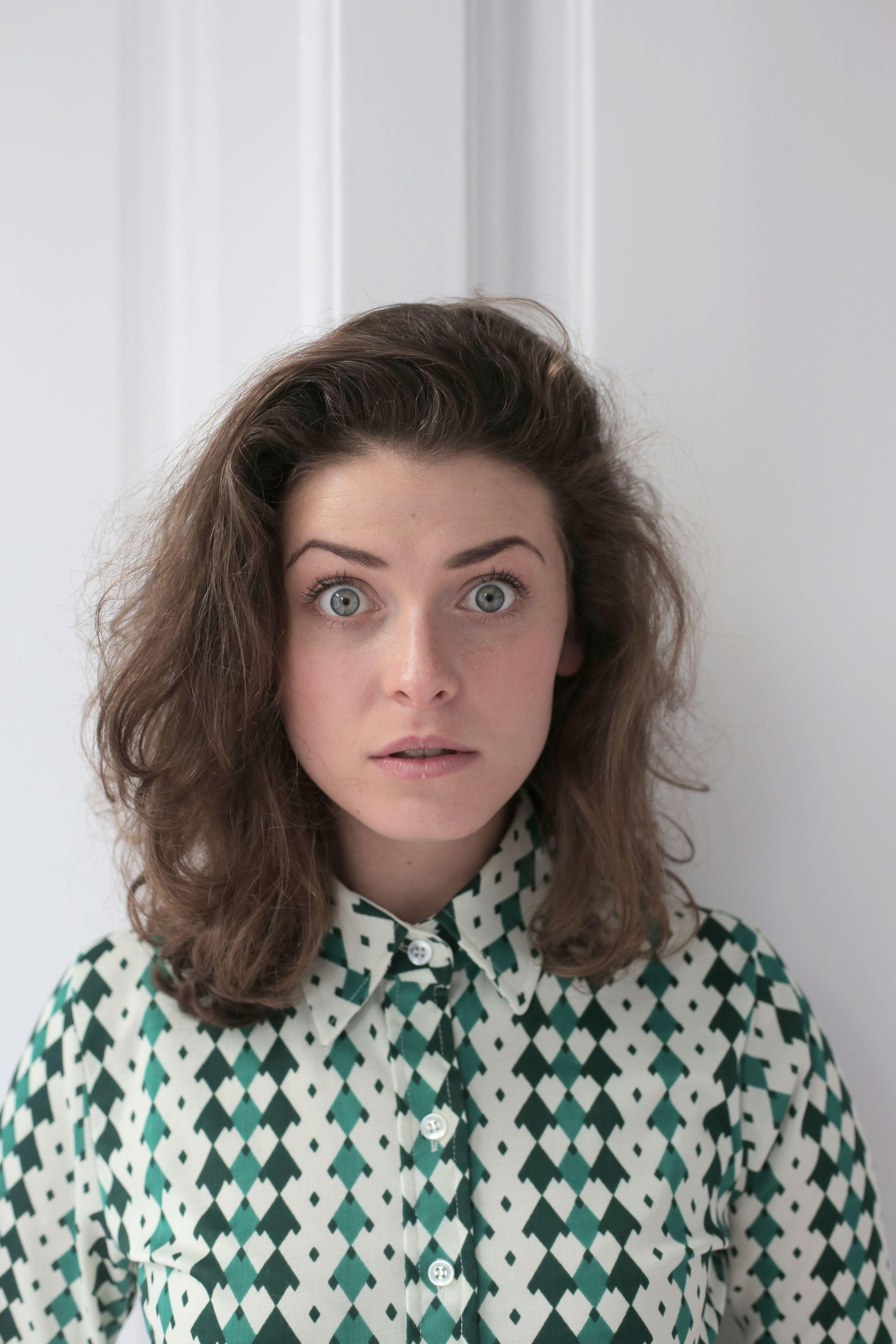 Shocked young lady with short wavy hair in knitted blouse standing near white wall in flat and looking at camera with wide open eyes