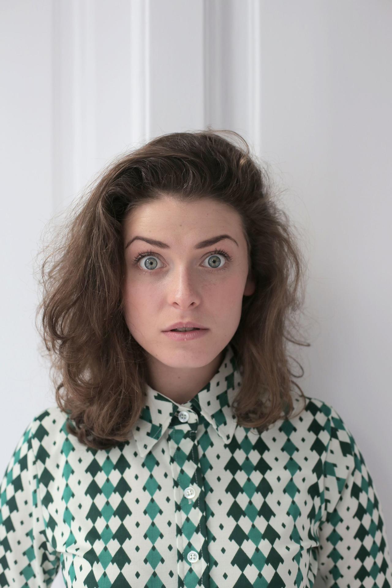 Shocked young lady with short wavy hair in knitted blouse standing near white wall in flat and looking at camera with wide open eyes