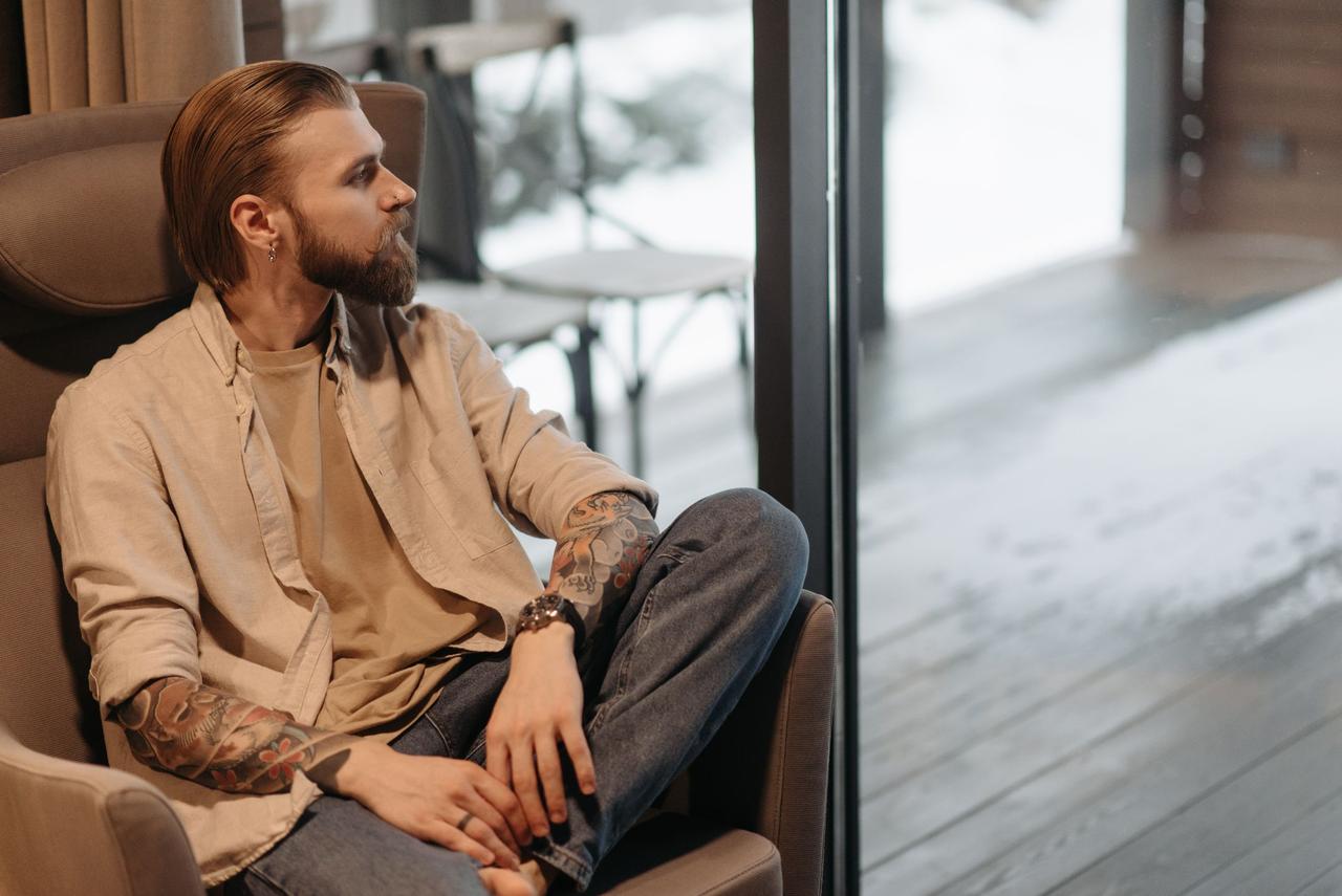 A bearded, tattooed man sits pensively, looking out a window on a winter day.