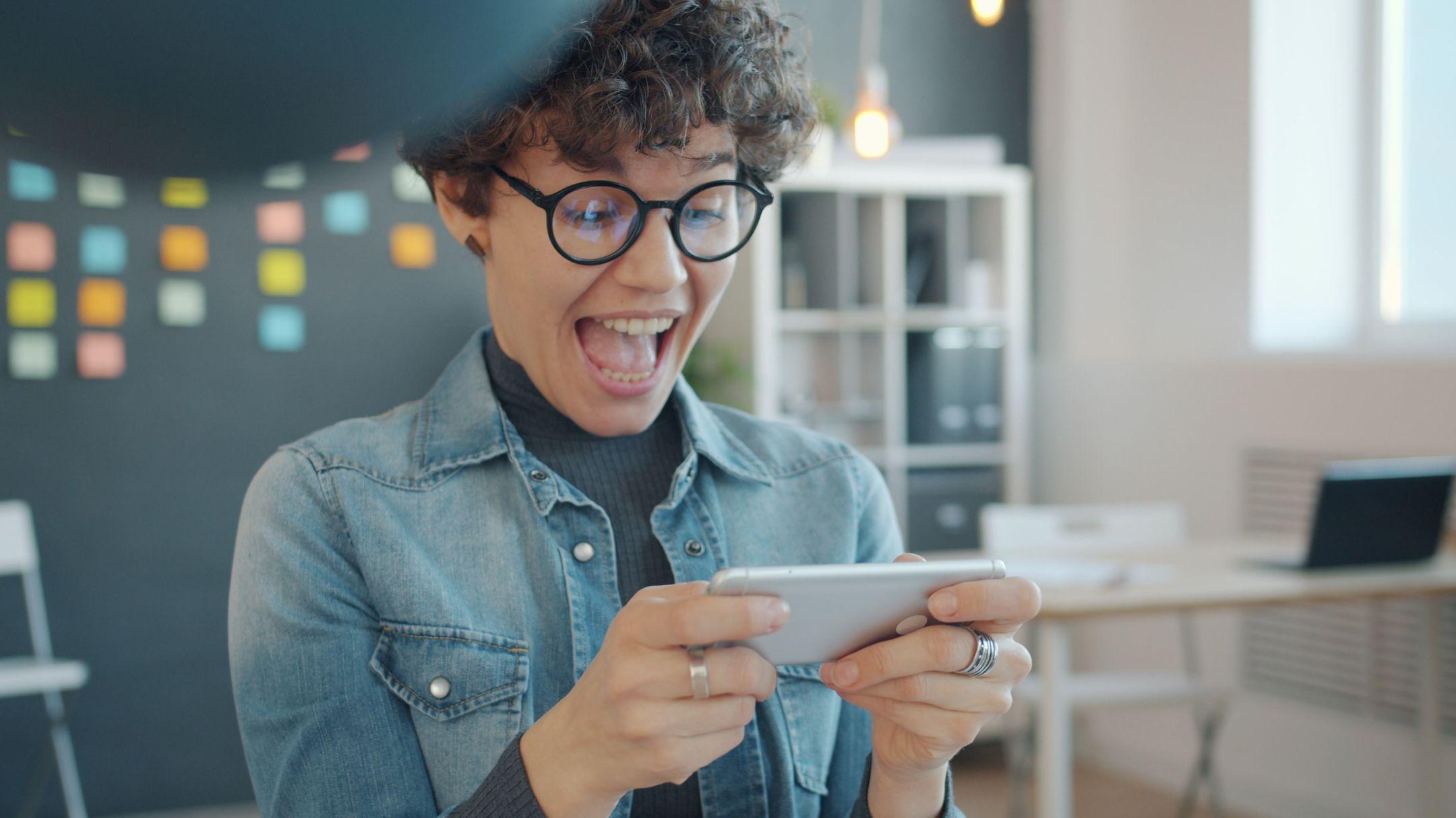 A young woman reacts with excitement while playing a mobile game indoors.