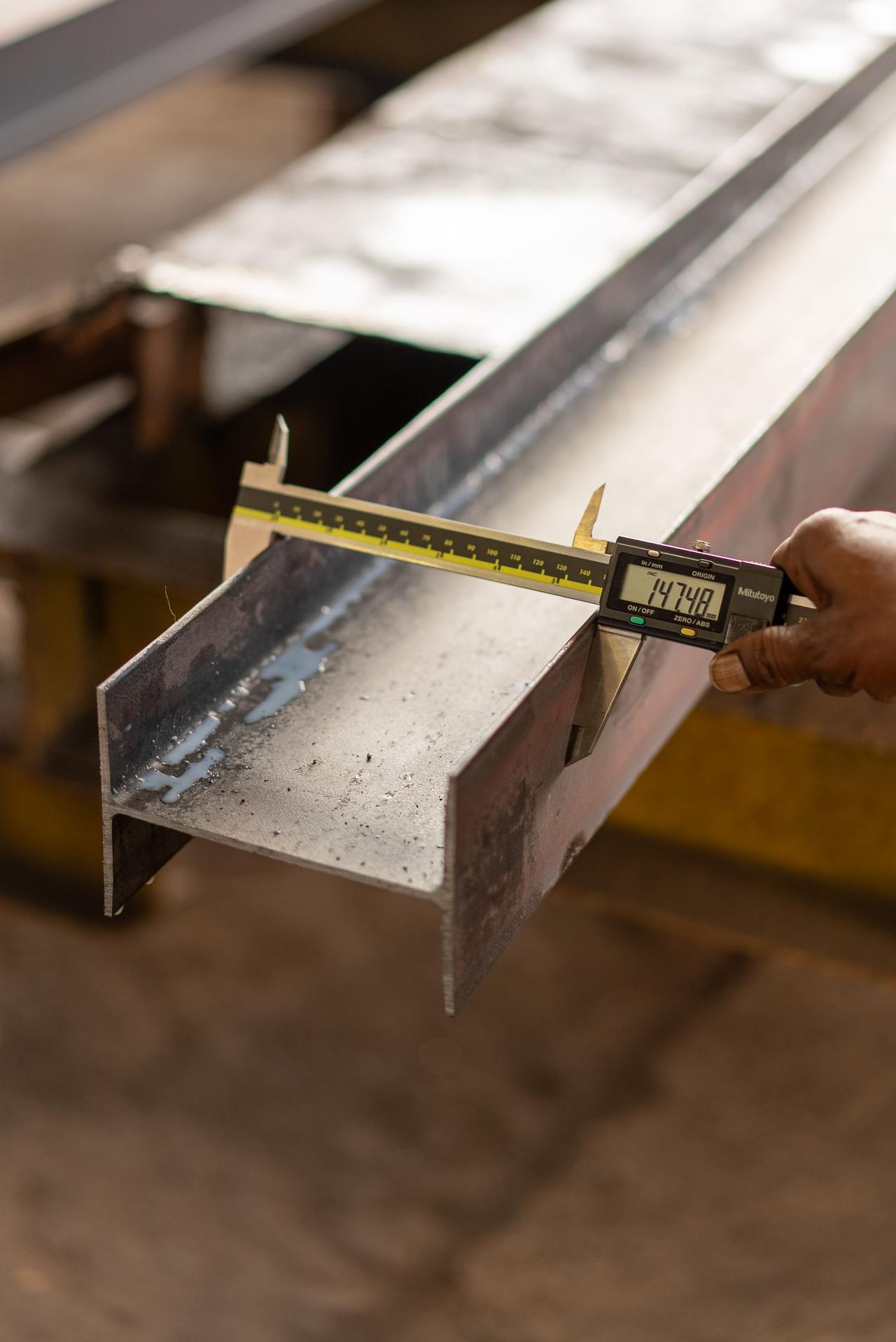 Close-up of a hand measuring a steel beam using a digital caliper in a workshop.