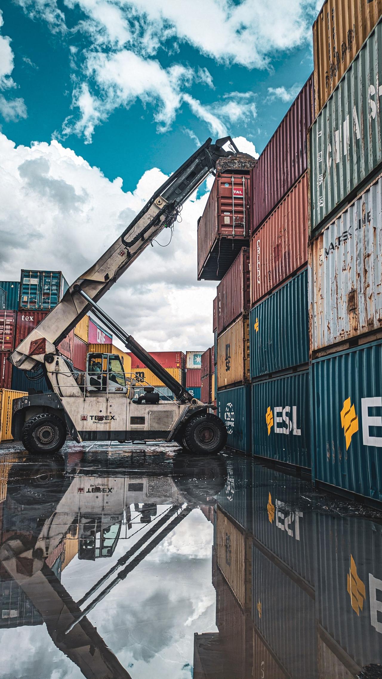 A crane loader stacking colorful shipping containers at a port with reflection in water.