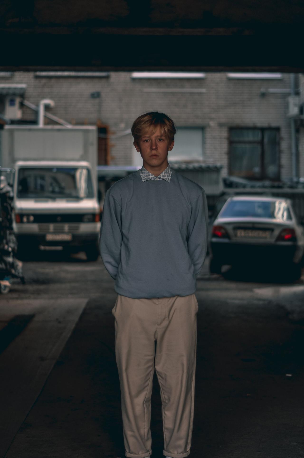 A young man stands in an urban alley, wearing a sweater and chinos. Moody atmosphere.