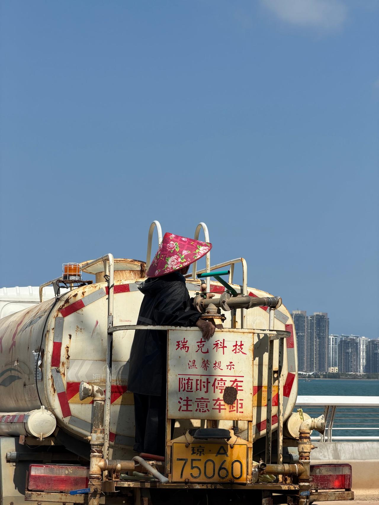 Worker in colorful hat stands on water tanker against city skyline under clear blue sky.