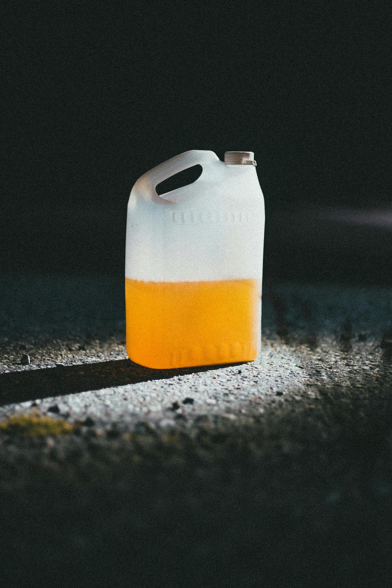 A plastic canister half-filled with orange liquid on a gravel floor, highlighted by dramatic lighting.