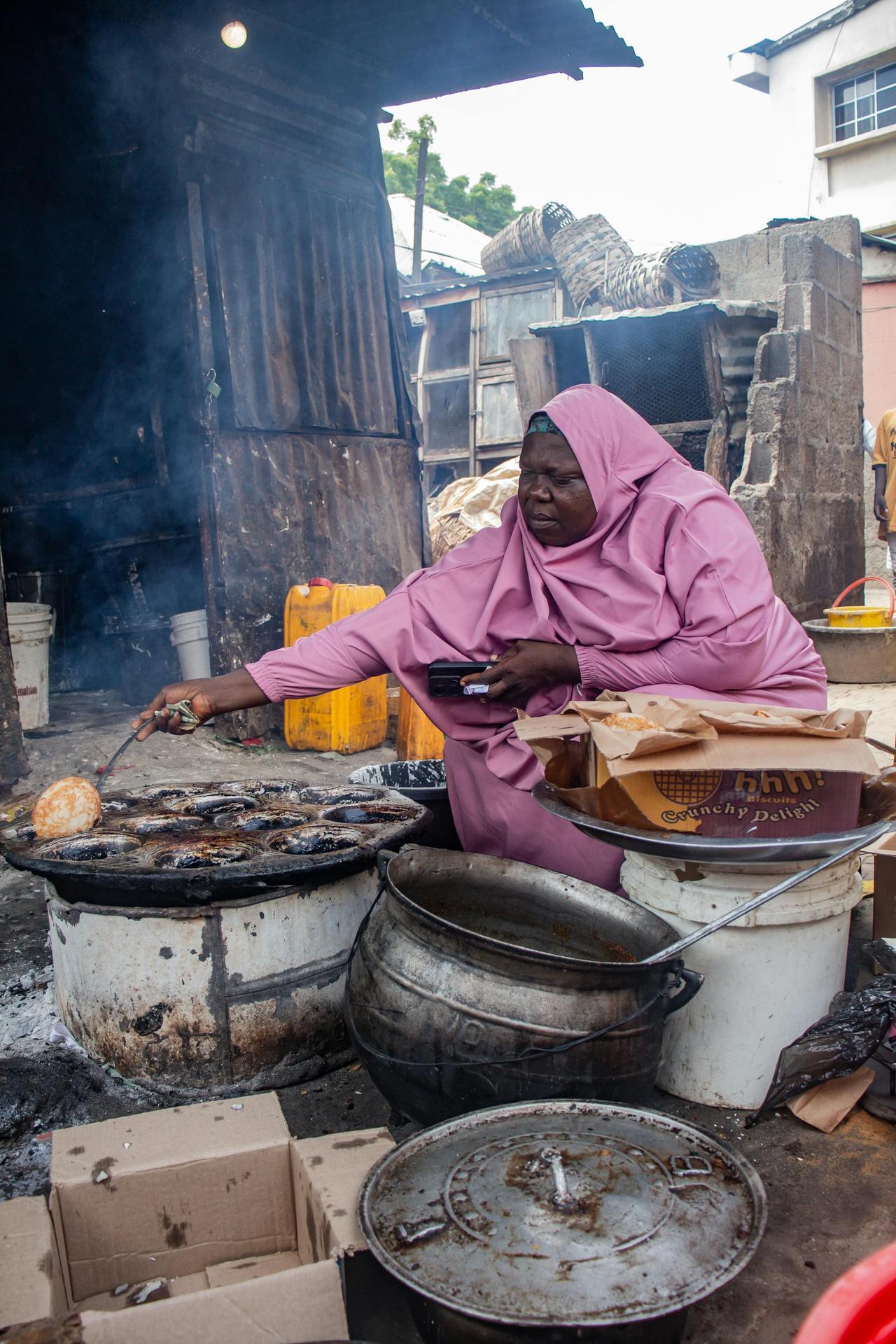 A Nigerian woman in pink attire cooking food on an open flame at a street market in Nigeria.