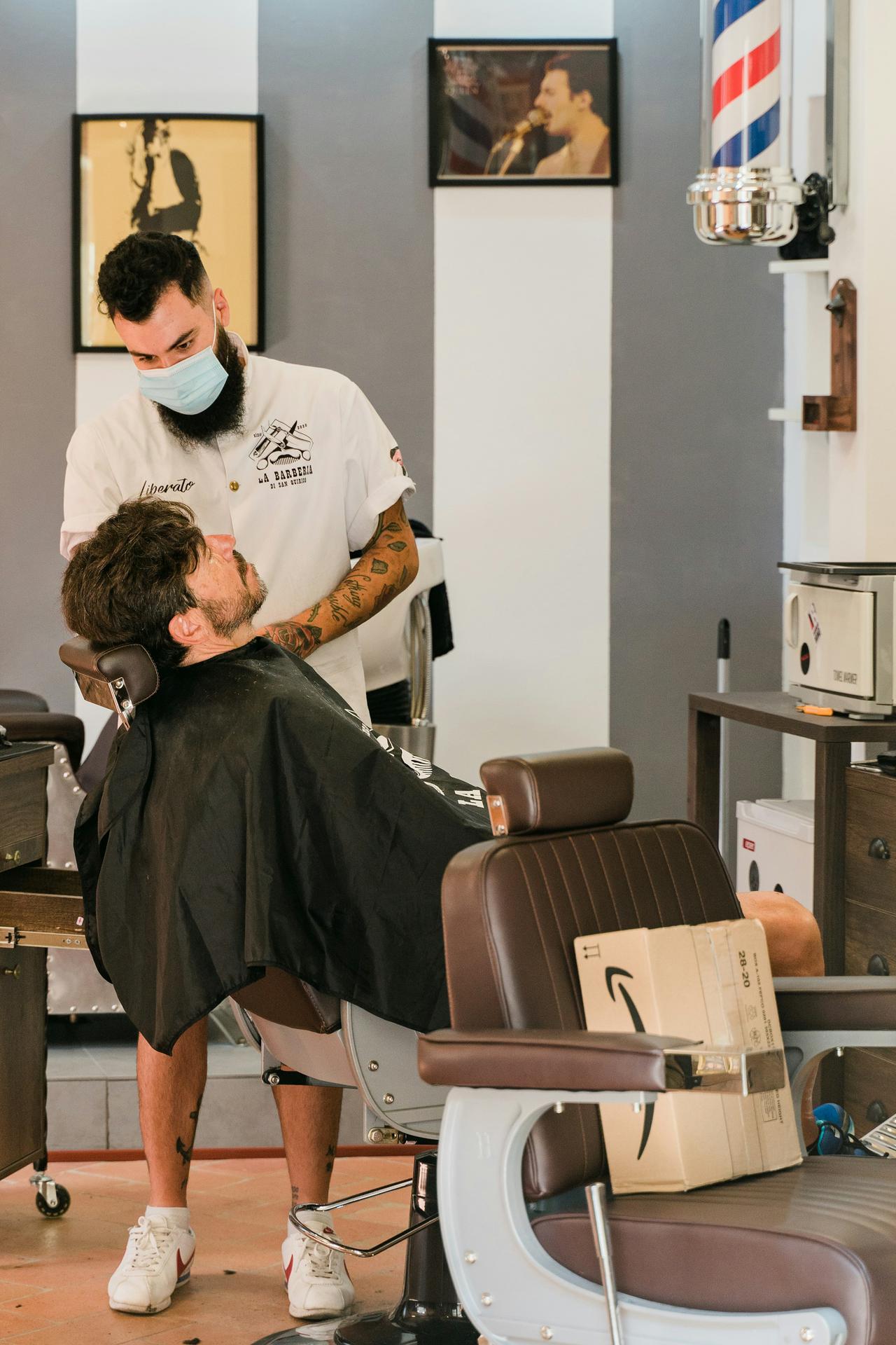 A bearded, tattooed young barber wearing a face mask trims a customer's hair in Italy