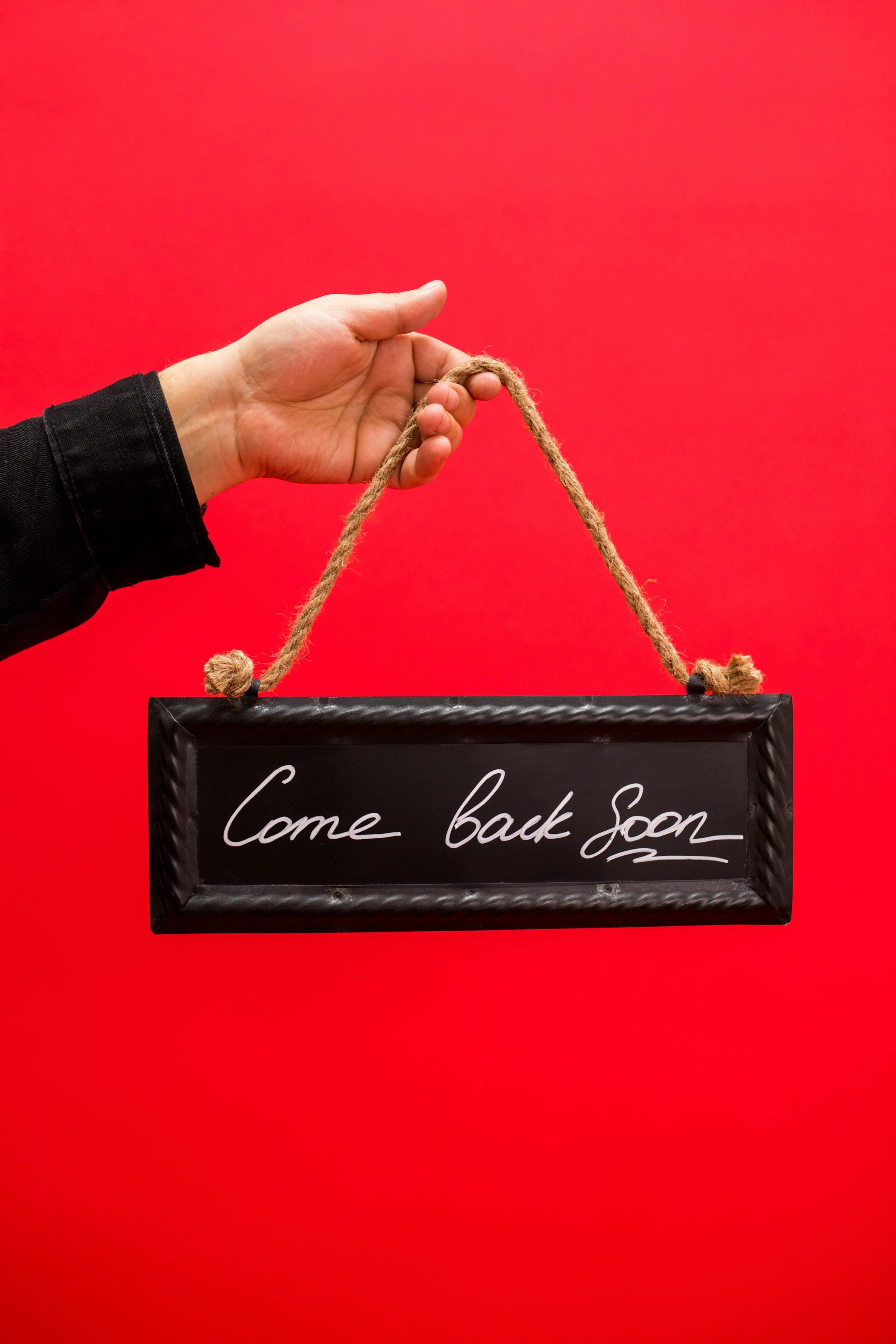 A hand holding a 'Come back soon' sign with a red background, perfect for farewell concepts.