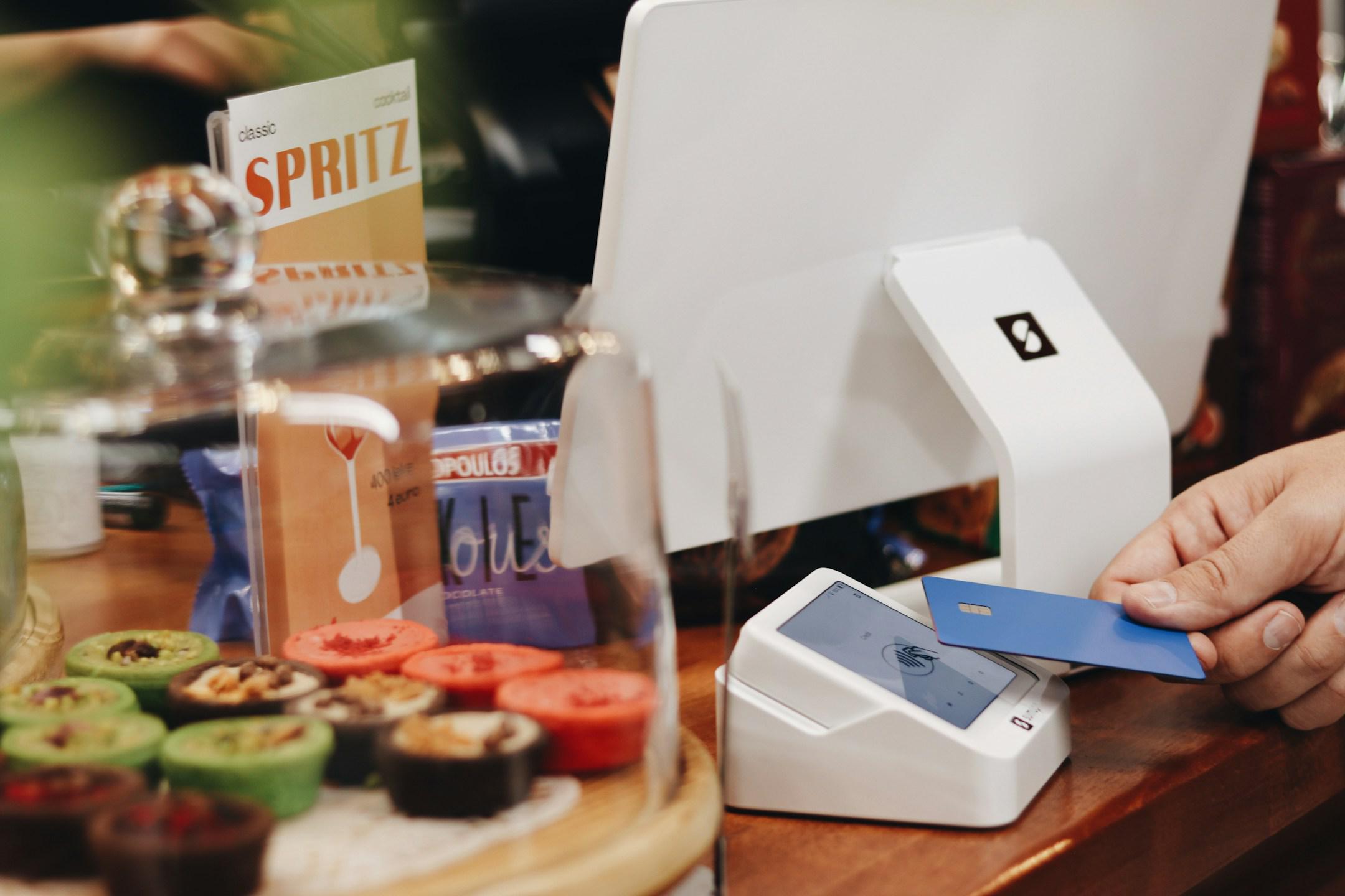 A customer is making a contactless payment using a blue card on a SumUp card reader in a cozy coffee shop or bakery. In the foreground, a variety of colorful desserts are displayed under a glass dome, adding to the inviting atmosphere of the store. A small sign featuring the word "Spritz" is also visible, highlighting products or promotions in the shop. The modern point of sale (POS) system suggests a seamless and efficient checkout experience for both the customer and the business.