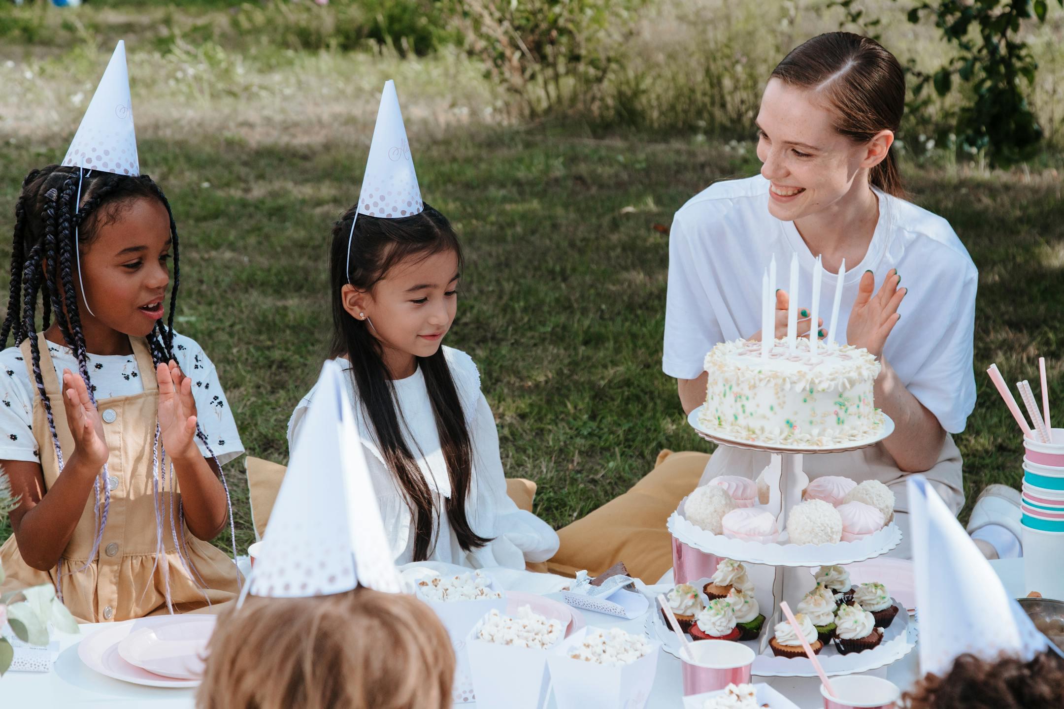 A delightful outdoor birthday party with kids and a woman celebrating with a cake and party hats.