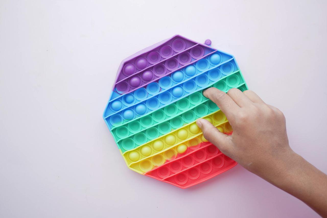 Close-up of a hand playing with a rainbow Pop It sensory toy on a white background.