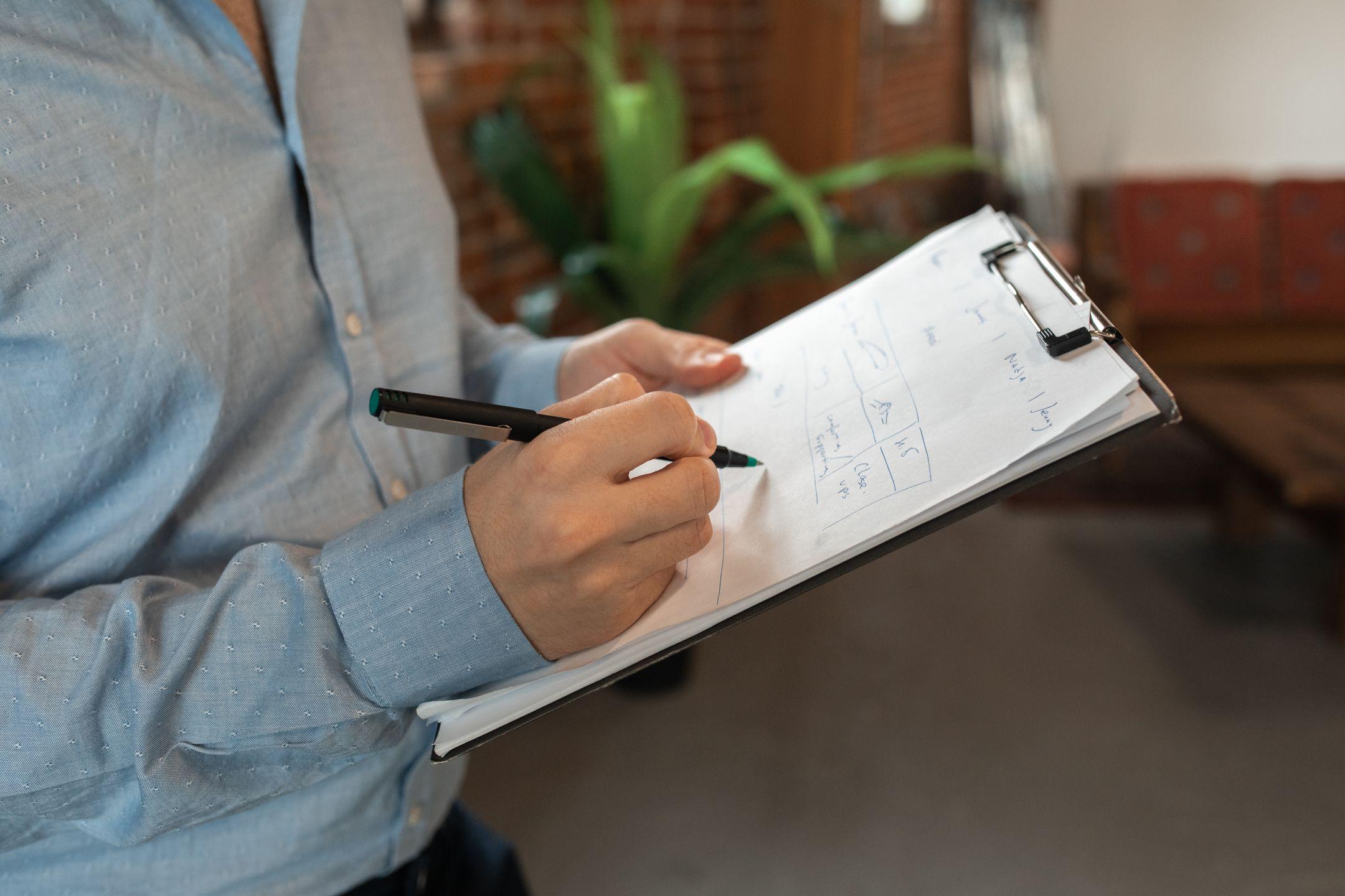 A person in blue shirt writes with a pen on a clipboard indoors, showcasing focus and productivity.