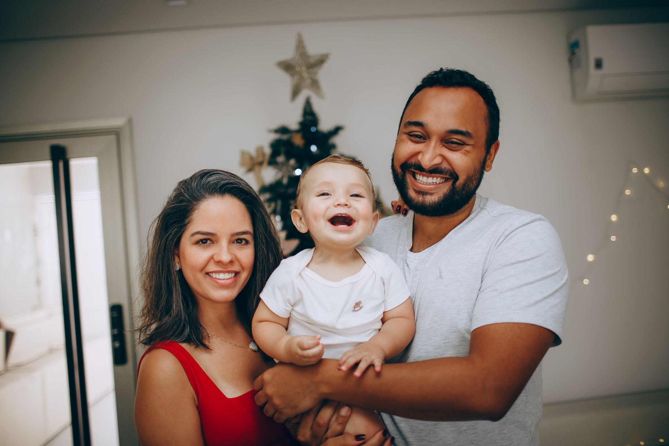A joyful family of three smiling in front of a decorated Christmas tree indoors.