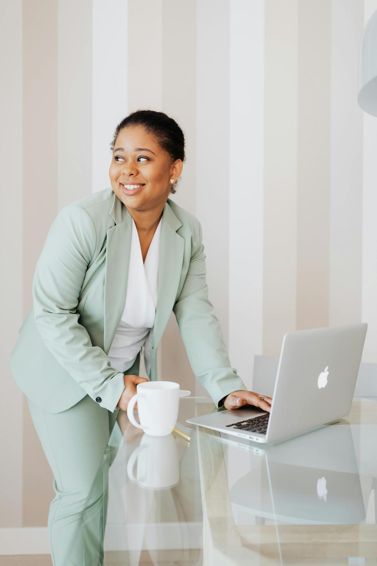 Smiling woman in a mint suit working on a laptop indoors, holding a coffee cup.