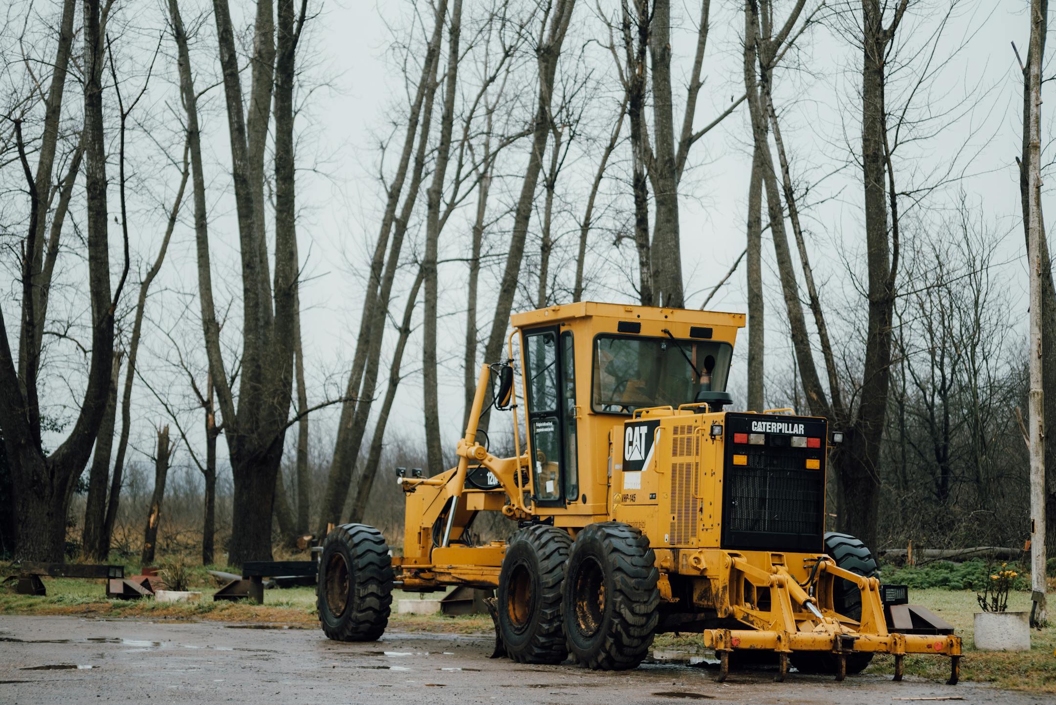 A yellow Caterpillar grader parked in a forest clearing during a cloudy day.
