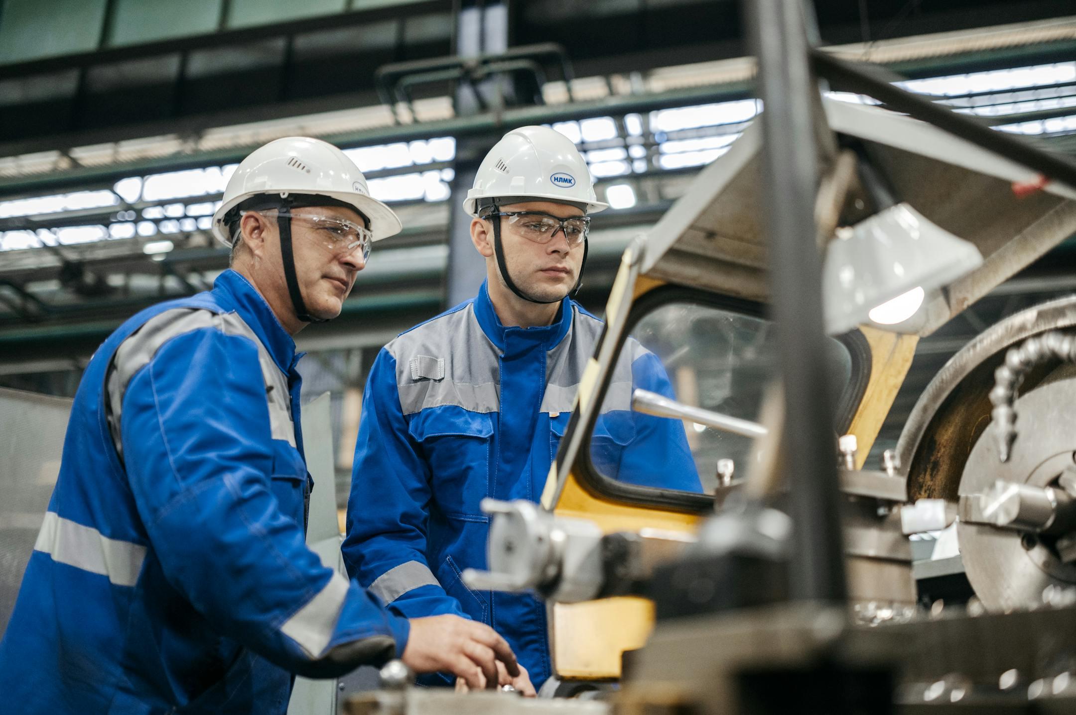 Two industrial workers in protective gear operate machinery in a factory setting.