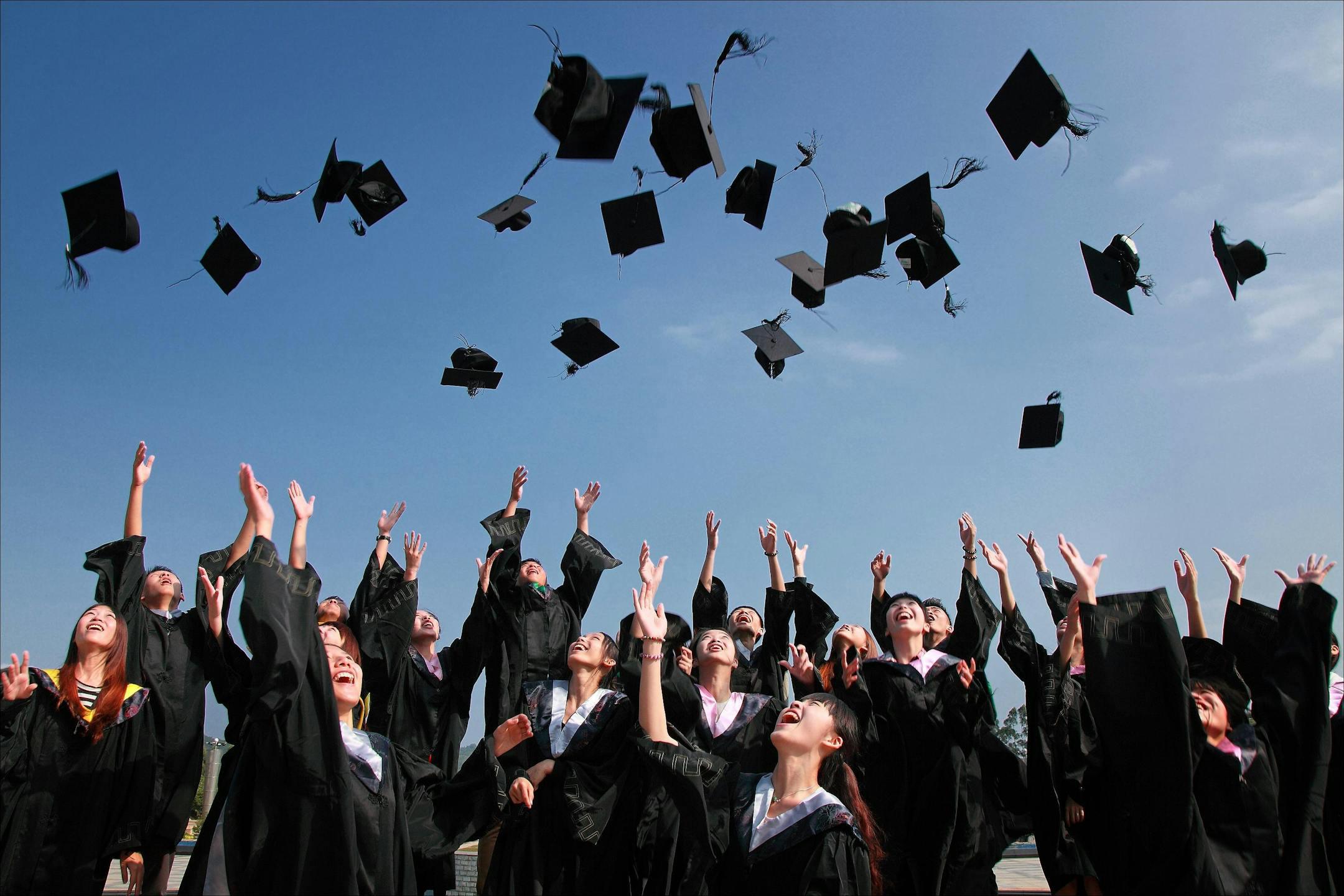 Group of graduates celebrating by throwing caps in the air during a sunny day. This level of education is made possible by a top business consulting firm in Michigan.