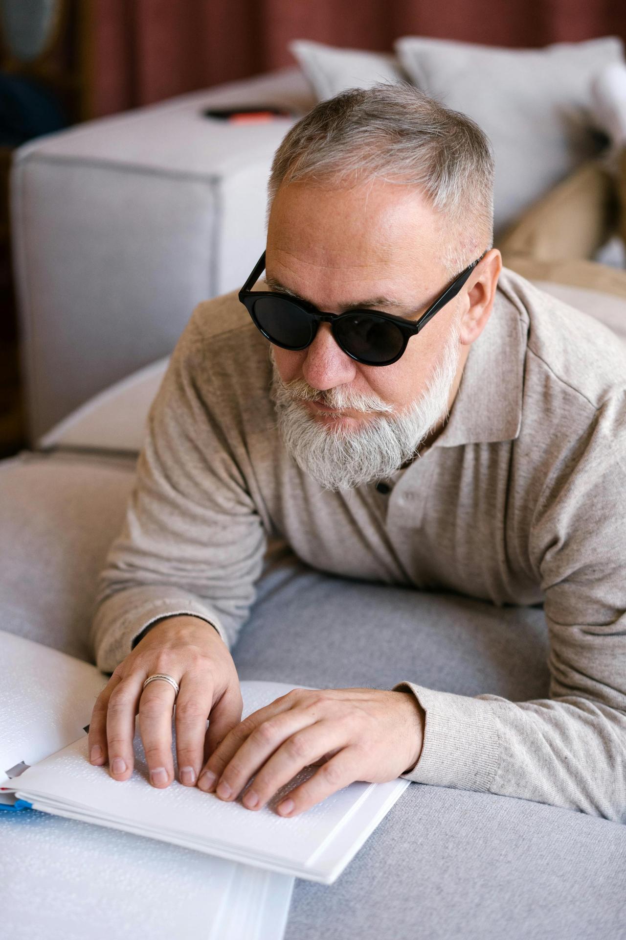 Elderly man wearing sunglasses reading a Braille book indoors for visual impairment awareness.
