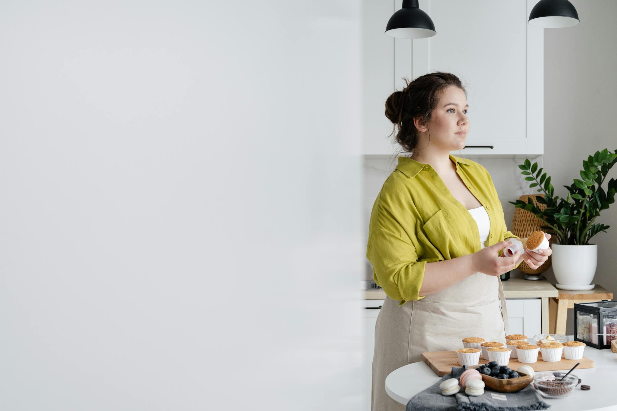 Female cook standing near table and looking away while preparing tasty cupcakes in light kitchen