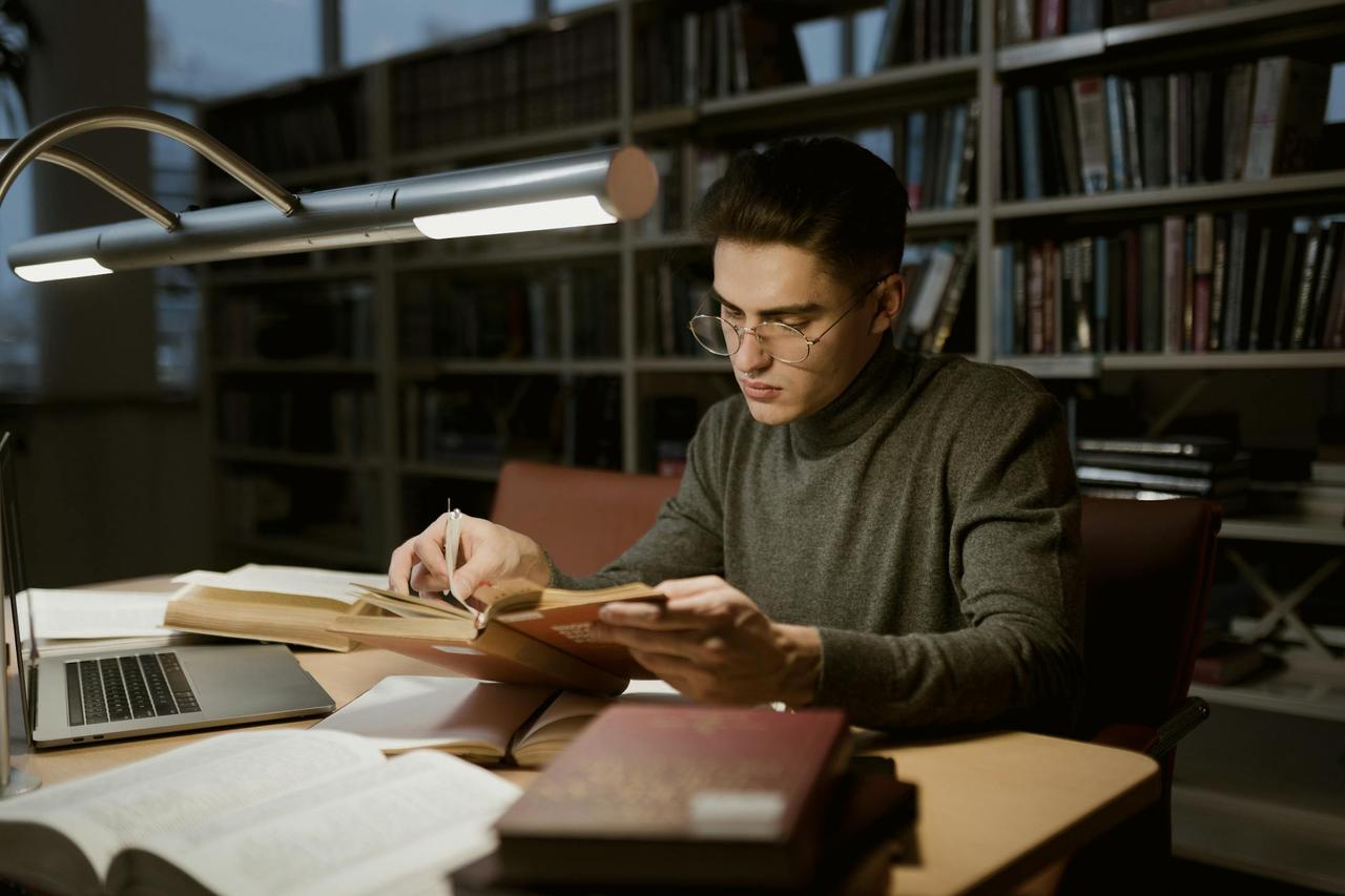 A young man in a sweater reads books in a library, surrounded by books and a laptop.