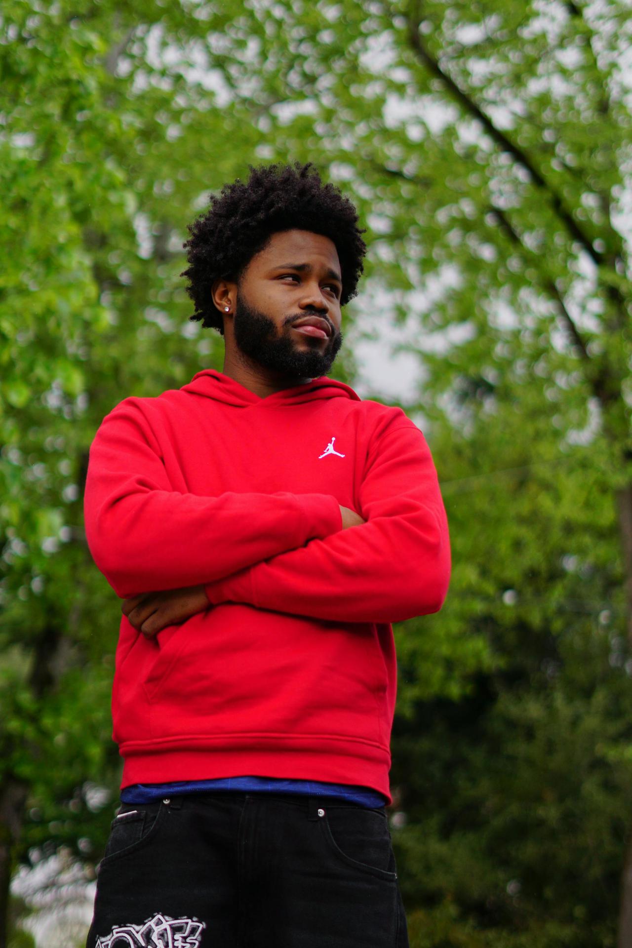 Portrait of a young man in a red hoodie, arms crossed, standing outdoors against a green background.