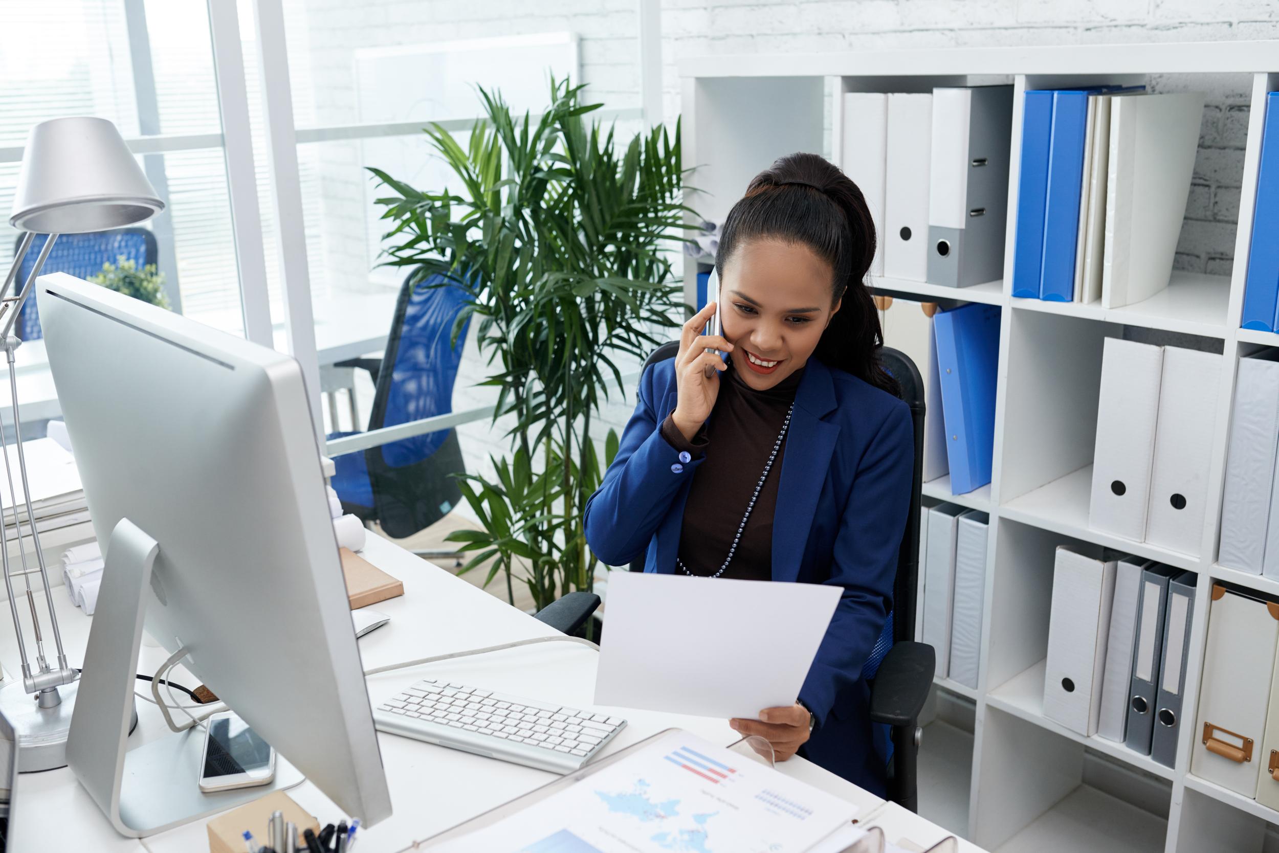 smiling-asian-woman-sitting-desk-office-looking-document-talking-mobile-phone.jpg