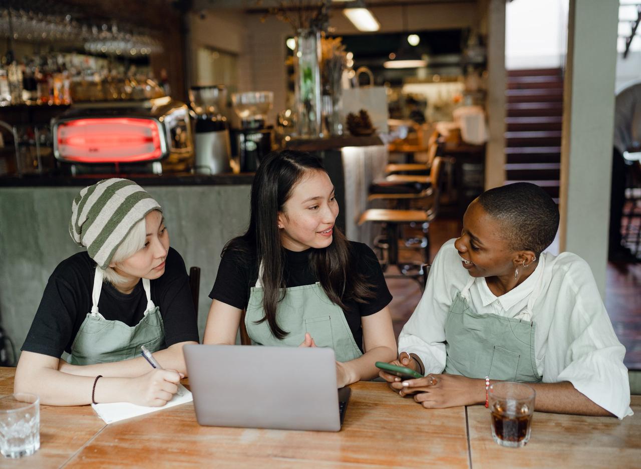 Positive young multiethnic waitresses in aprons sitting at wooden table in modern cafe and working on startup project on contemporary netbook