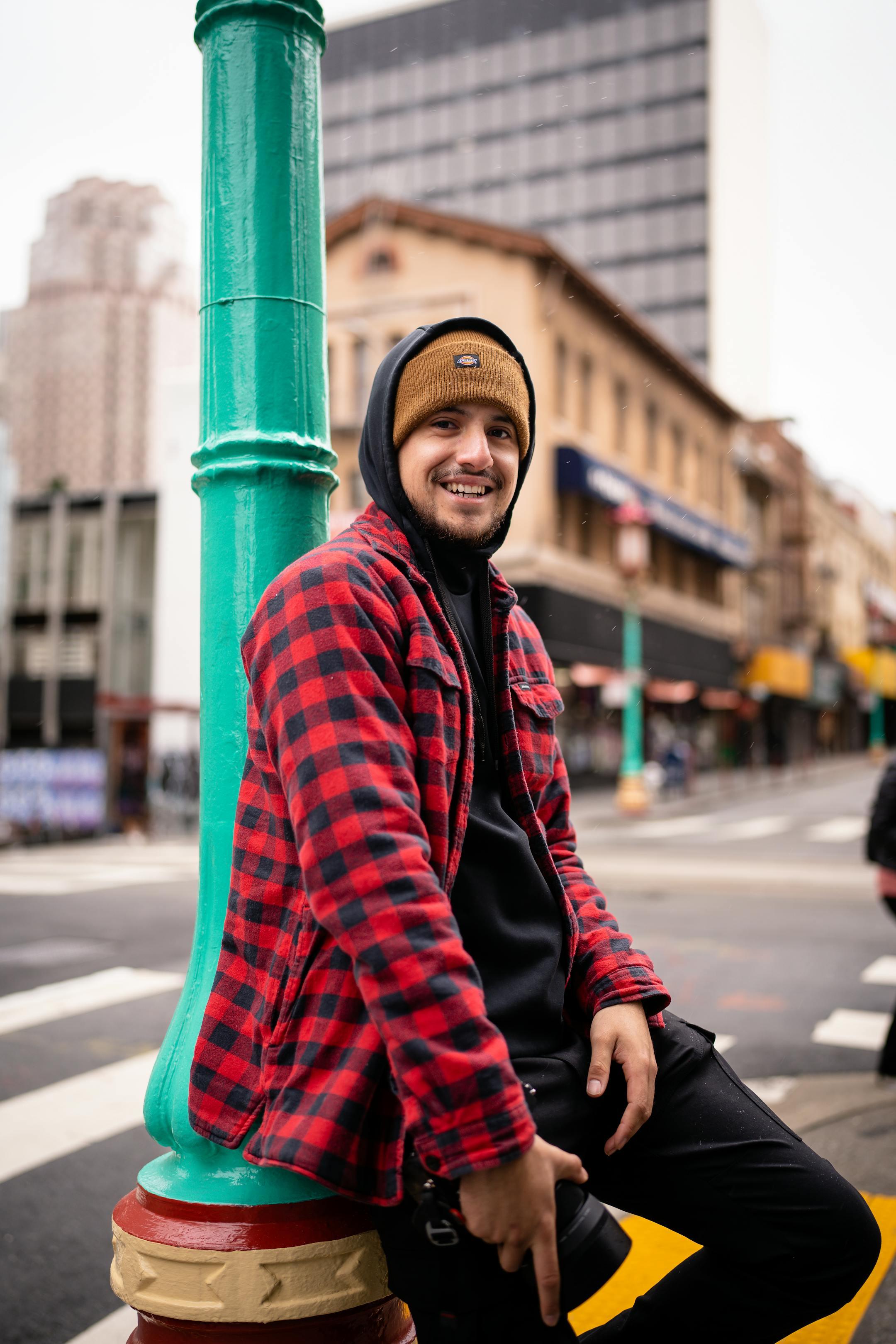 Smiling man in a red plaid jacket leaning on a colorful street pole in a city setting.