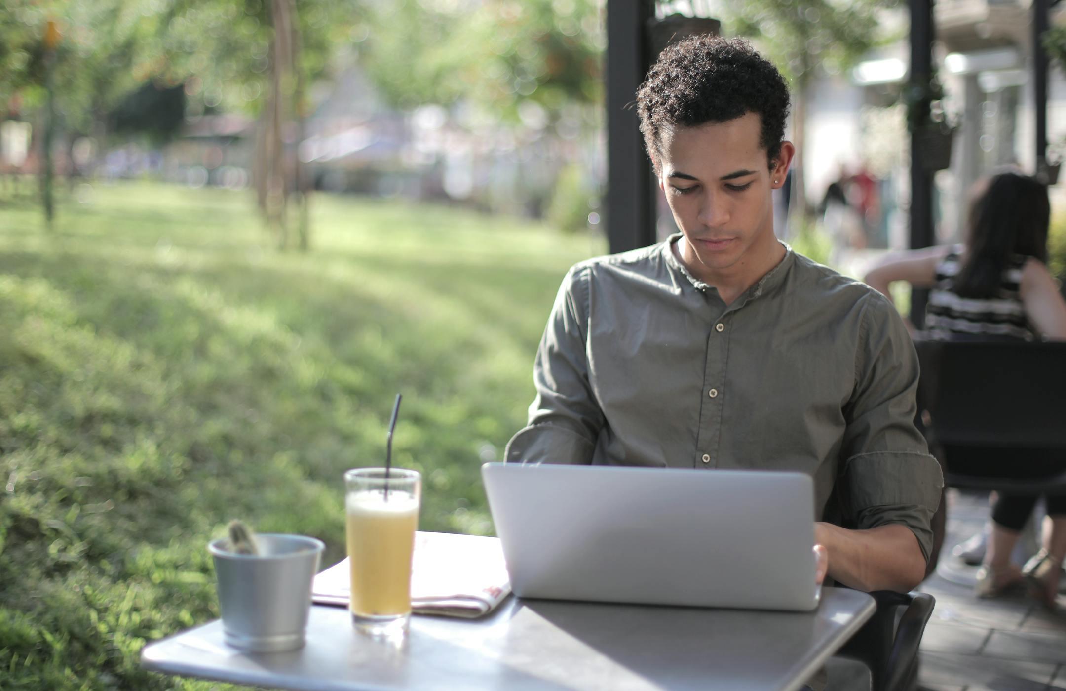 Young man working on a laptop outside in a casual and relaxed setting, enjoying a drink.