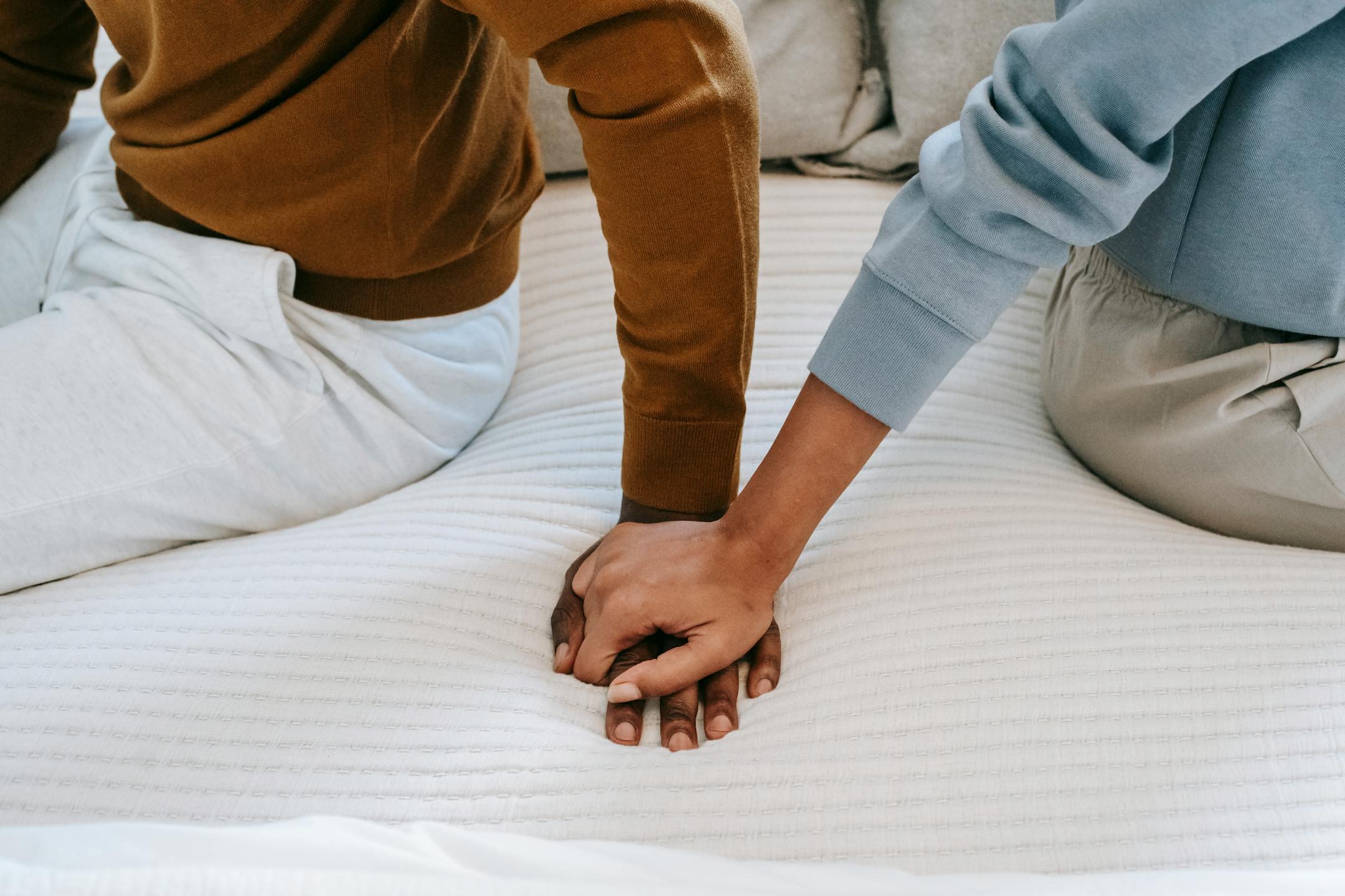 A couple sits closely on a bed holding hands, symbolizing connection.