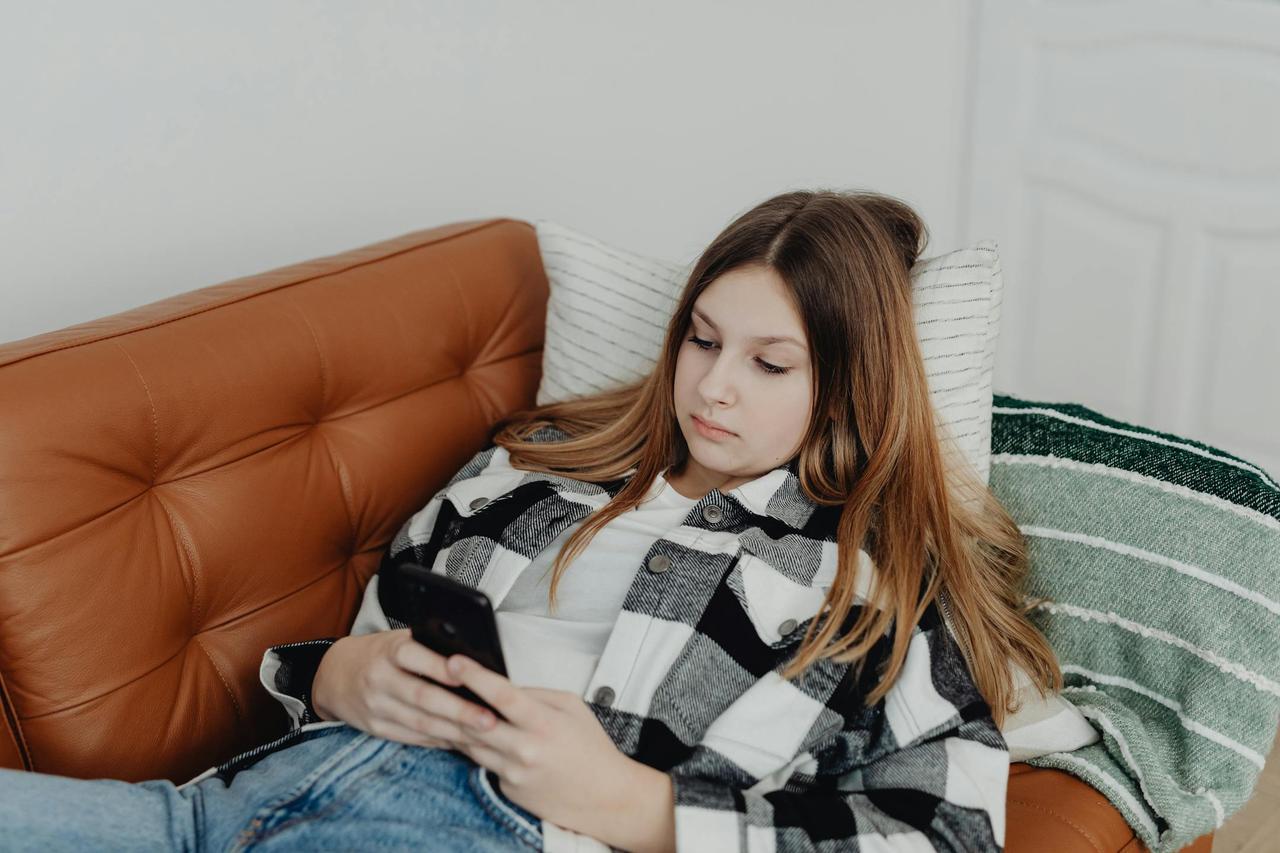 Teen girl lying on sofa using smartphone, enjoying casual indoor relaxation.
