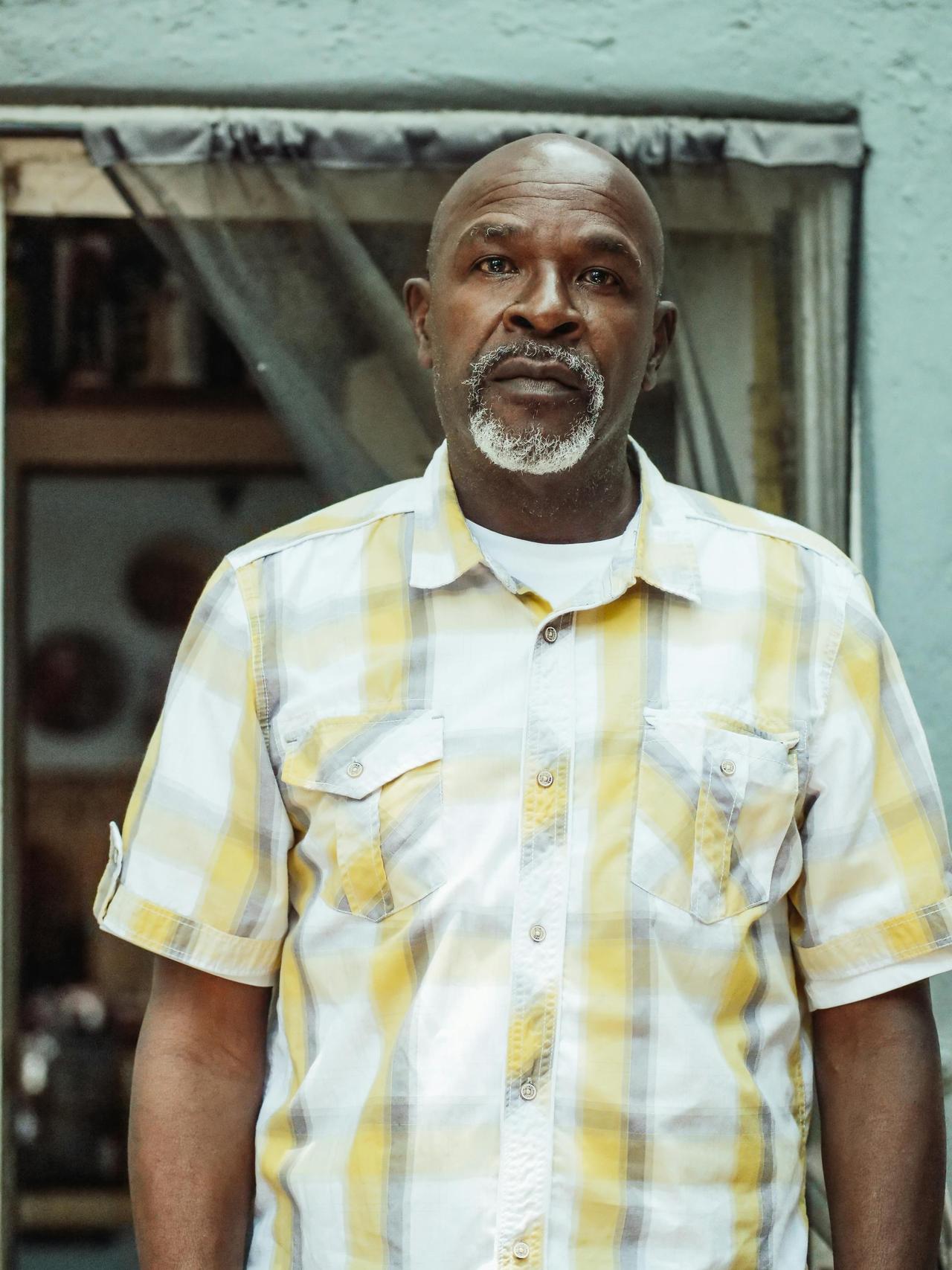 A senior man stands indoors wearing a checkered shirt, appearing contemplative.