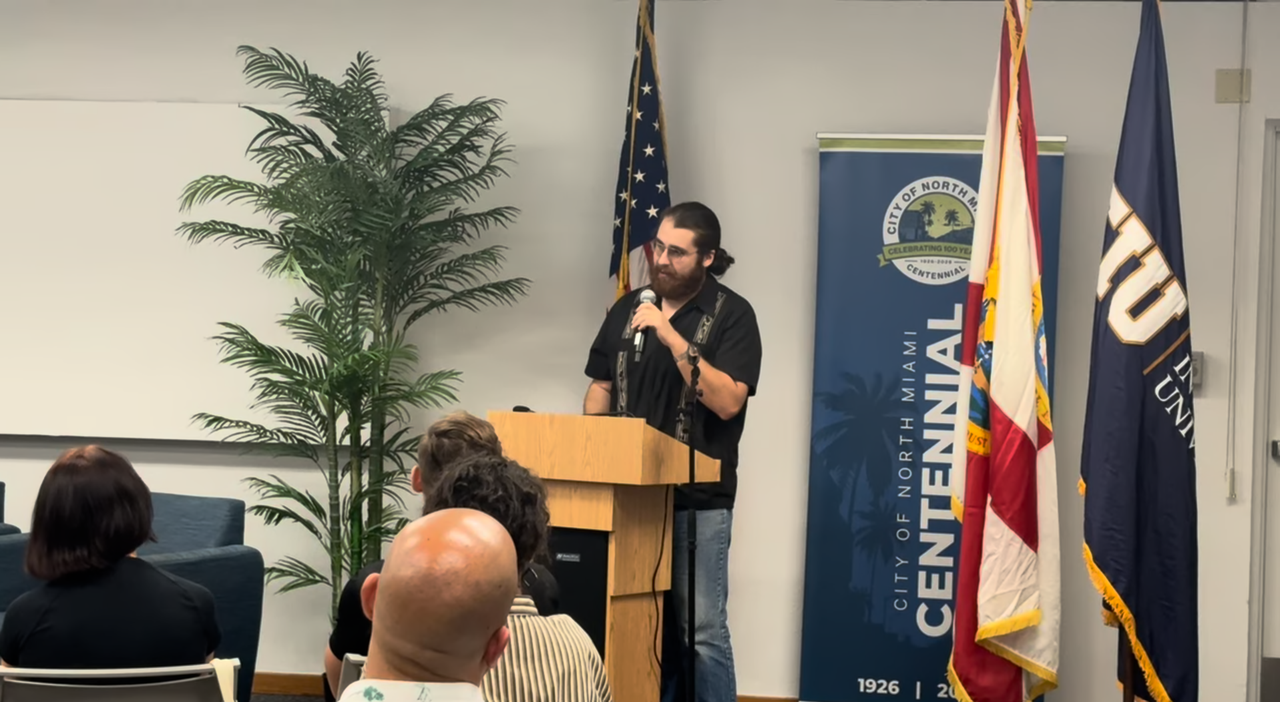 Chris reading at a lectern with the City of North Miami centennial banner and FIU flag beside him