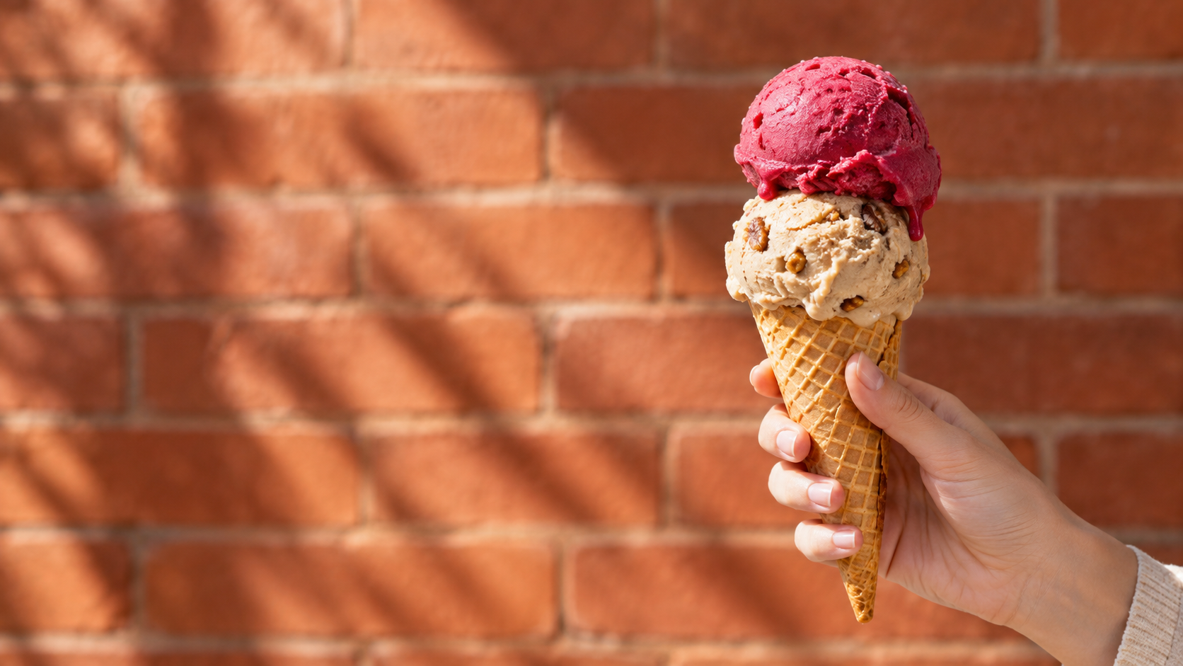 A hand holding a waffle cone with raspberry sorbet over maple walnut ice cream against a sunny brick wall