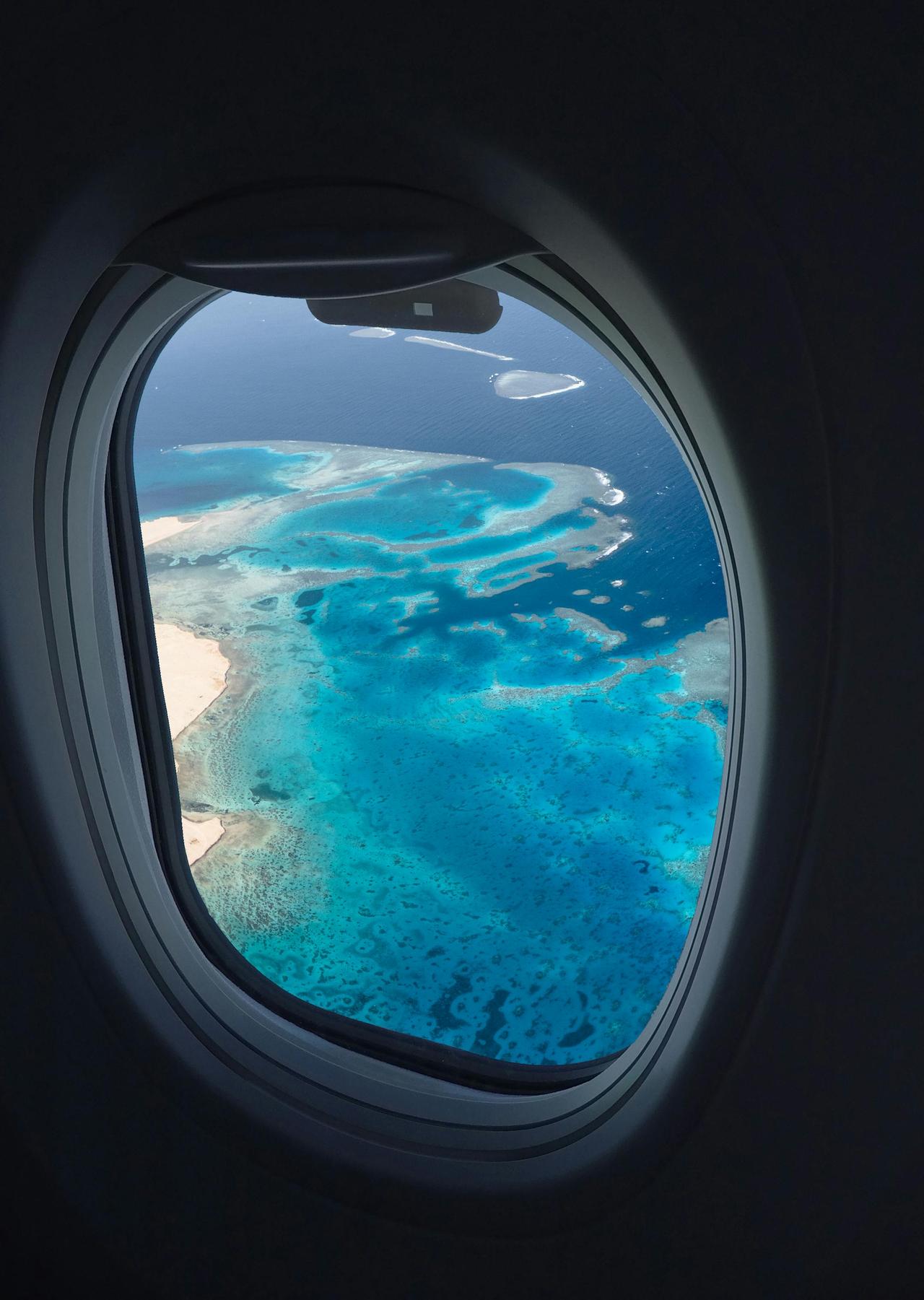 Breathtaking aerial view of ocean reefs through an airplane window, showcasing vibrant blues from above.