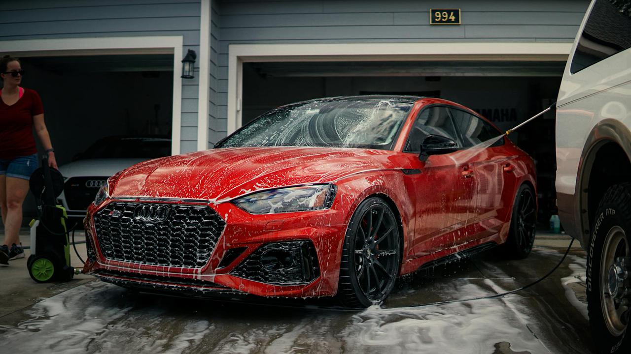 A red sports car is being cleaned with soap in a driveway, featuring a modern garage background.