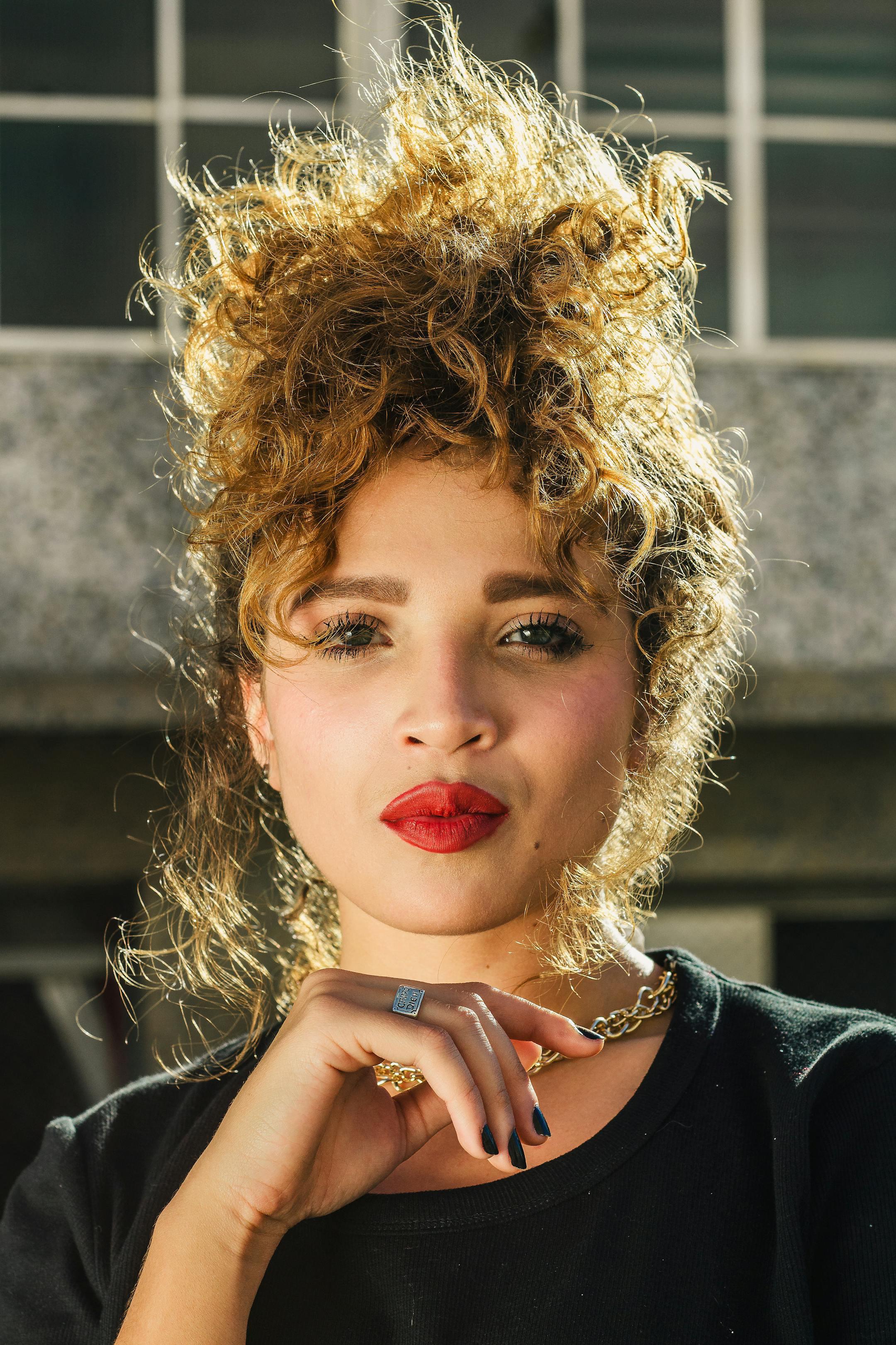 Portrait of a young woman with curly hair and red lips wearing a necklace outdoors.